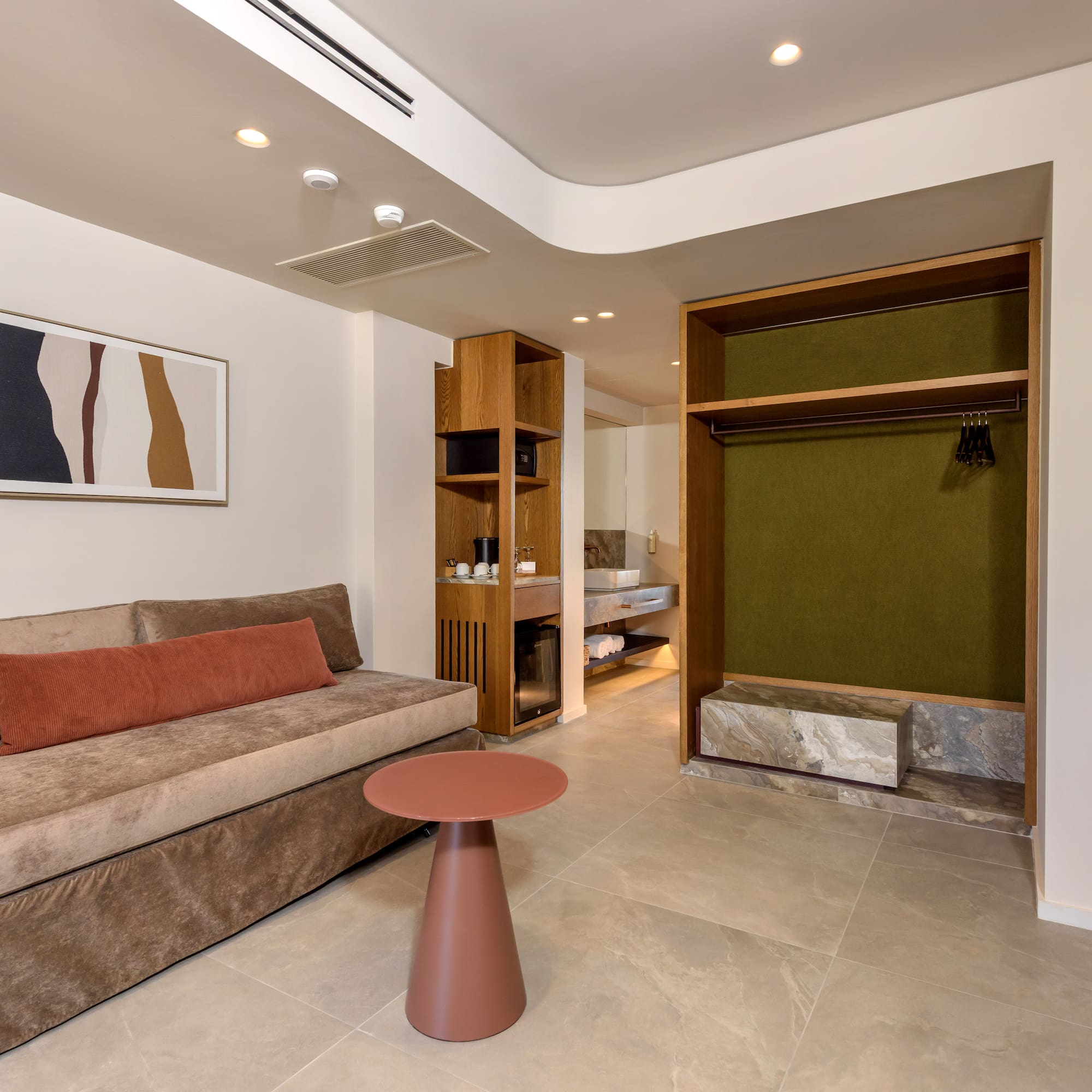 Modern hotel room with sofa, side table, kitchenette, and open wardrobe.
