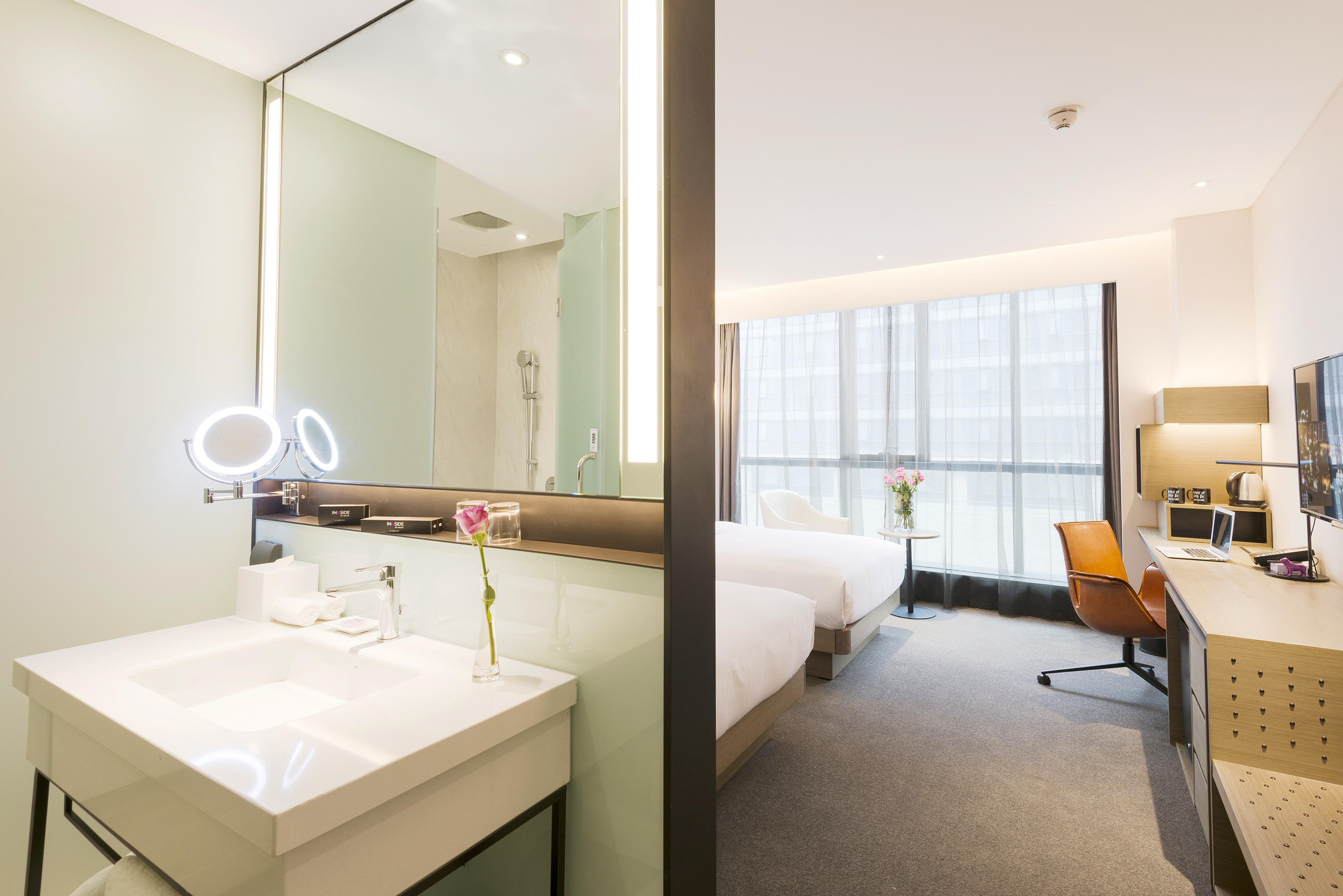 Bright hotel room with bathroom vanity, twin beds, and work desk.