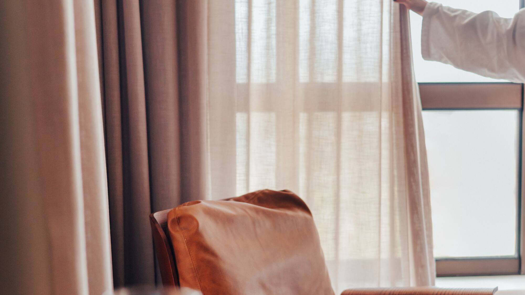 Woman opening bedroom curtains by a leather armchair, coffee, and book.