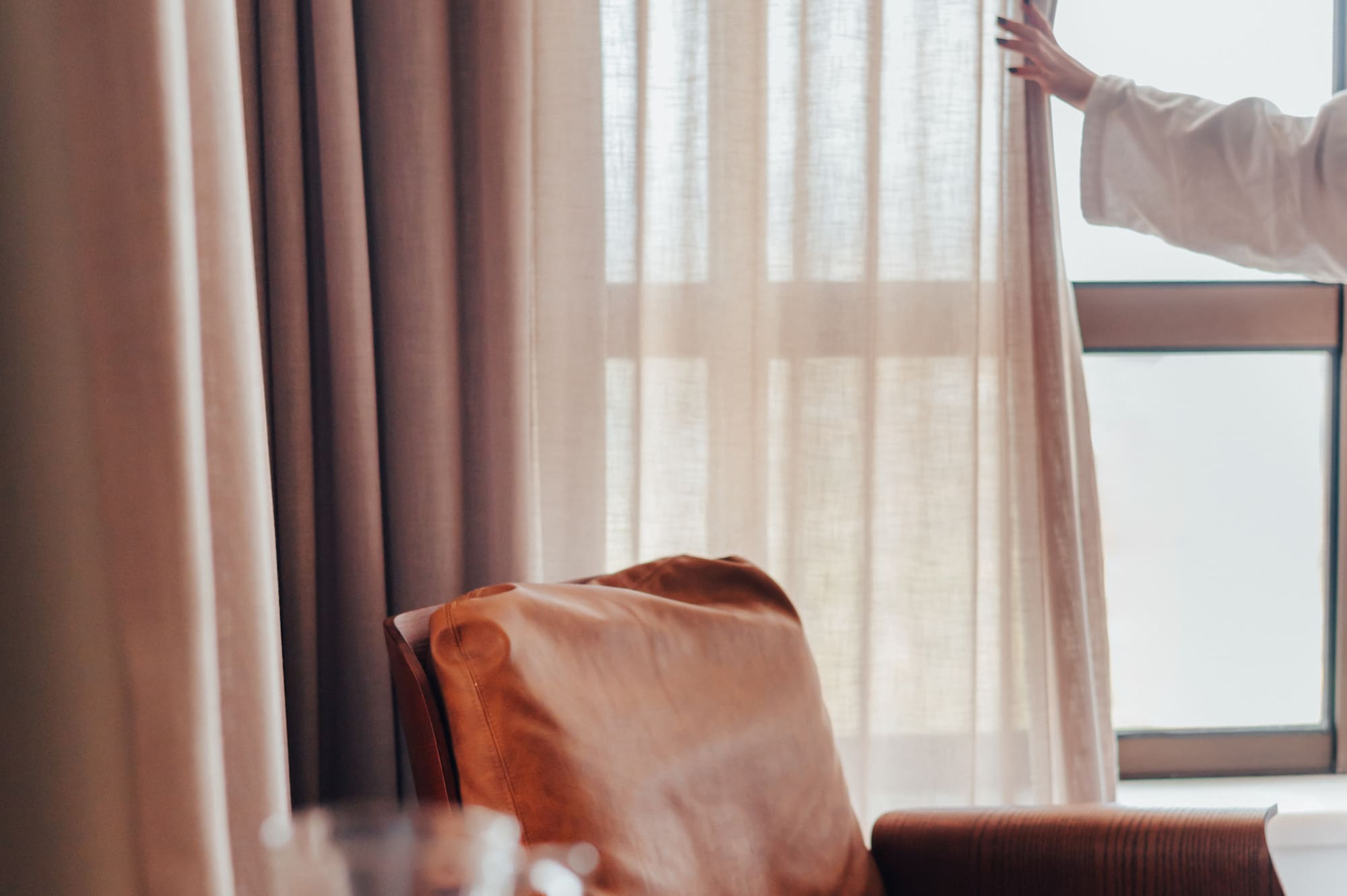 Woman opening bedroom curtains by a leather armchair, coffee, and book.