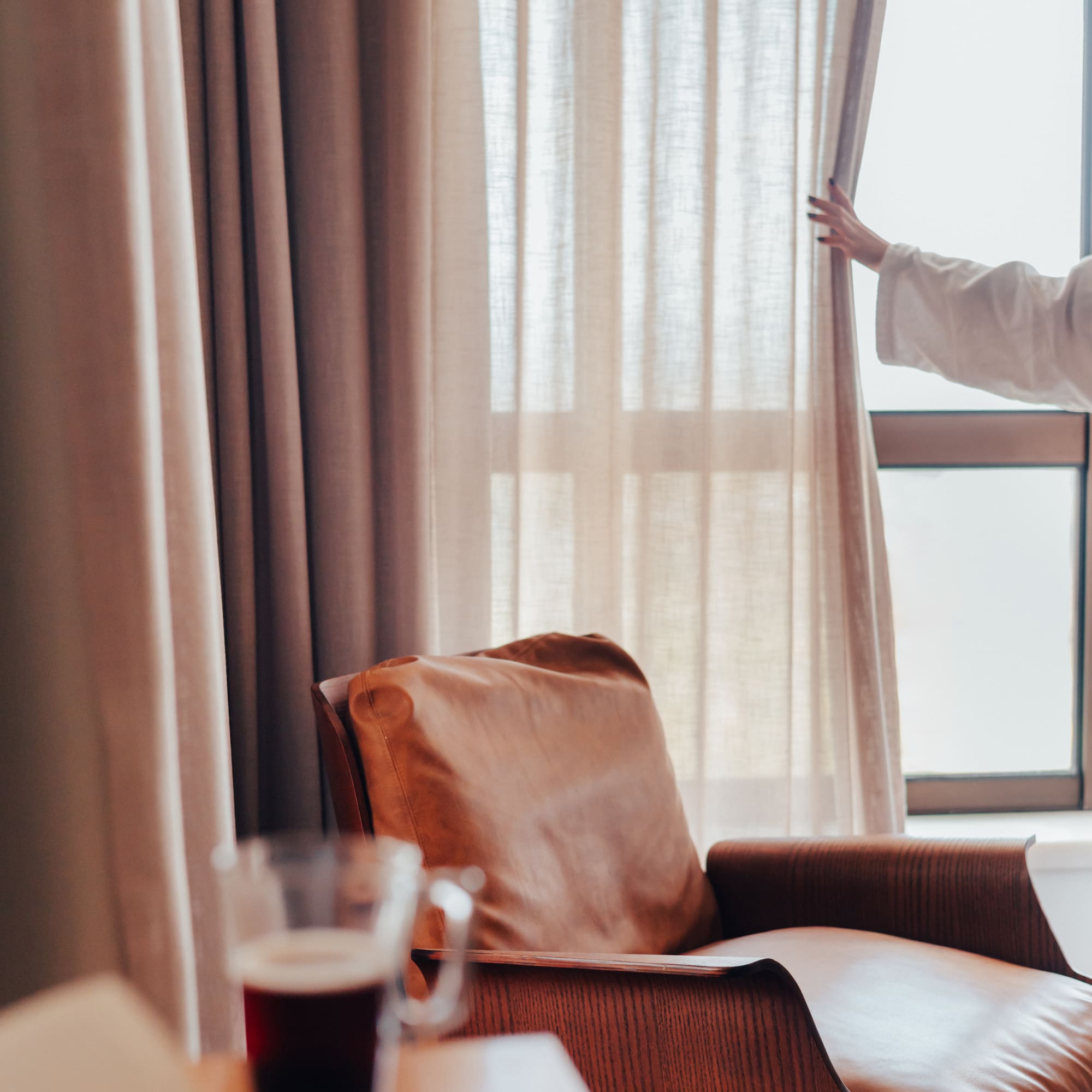 Woman opening bedroom curtains by a leather armchair, coffee, and book.