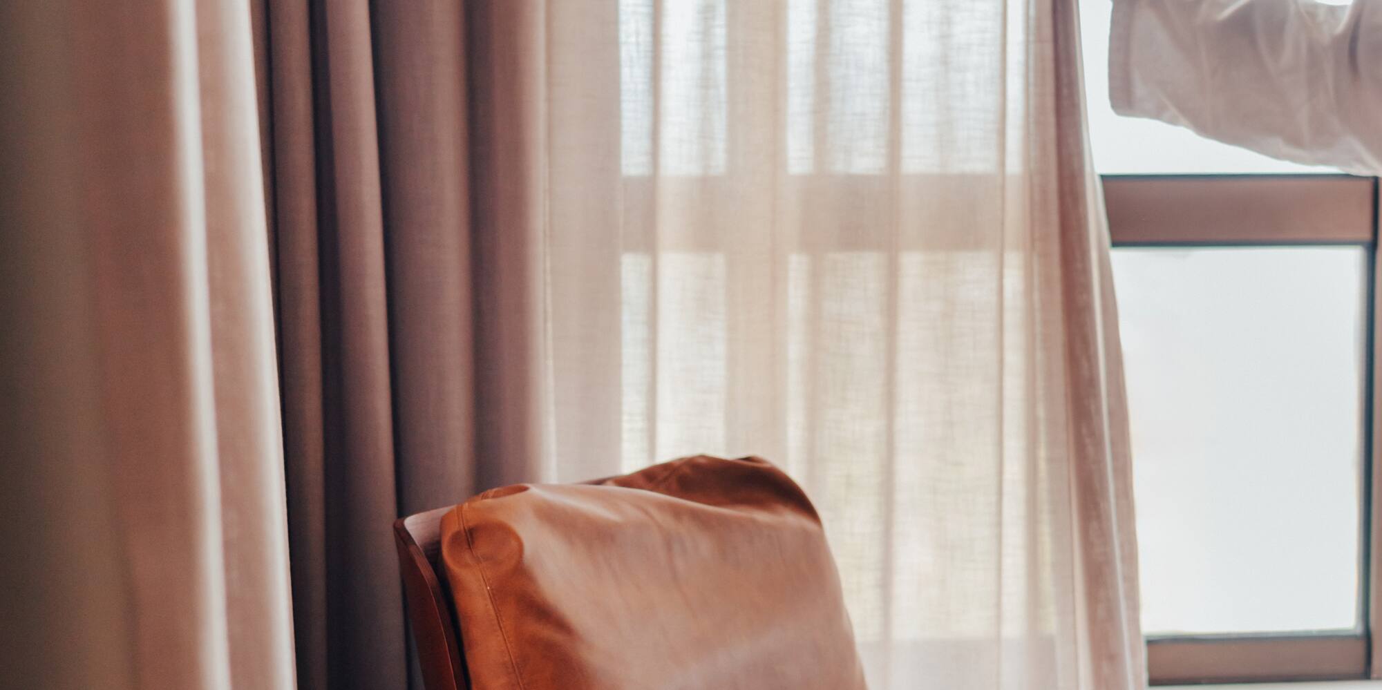 Woman opening bedroom curtains by a leather armchair, coffee, and book.