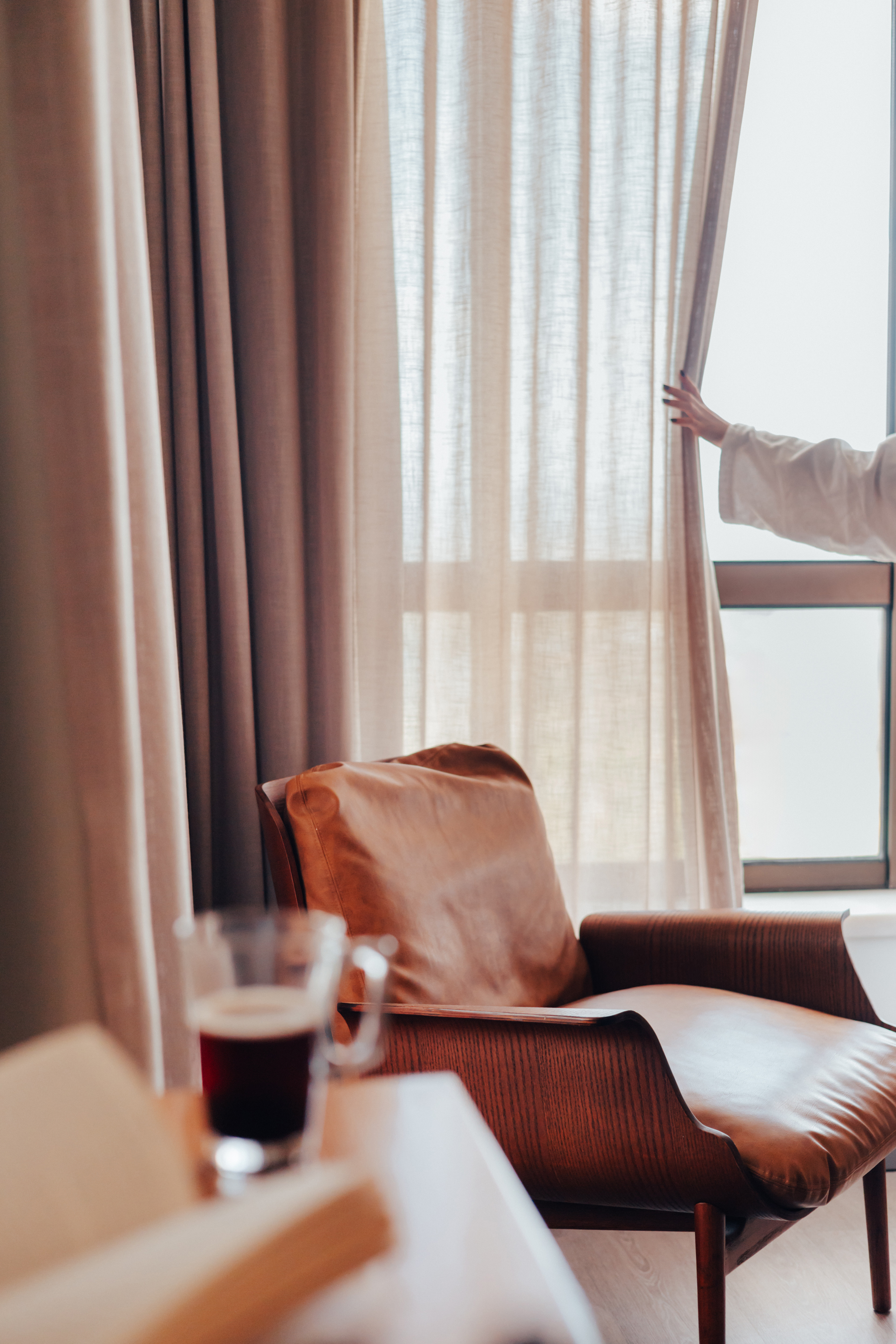 Woman opening bedroom curtains by a leather armchair, coffee, and book.