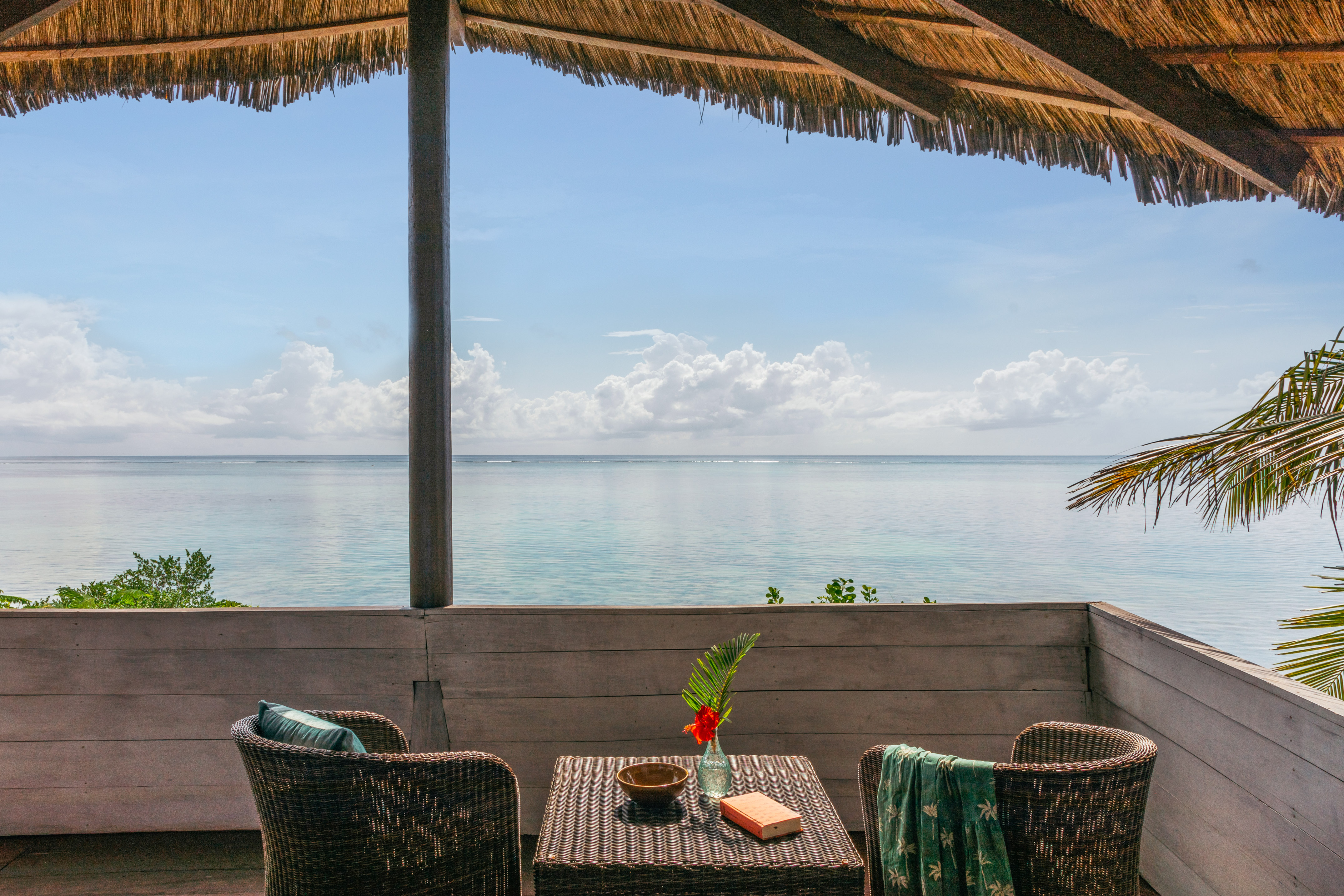 Tropical oceanfront balcony with rattan seating, table, and thatched roof.