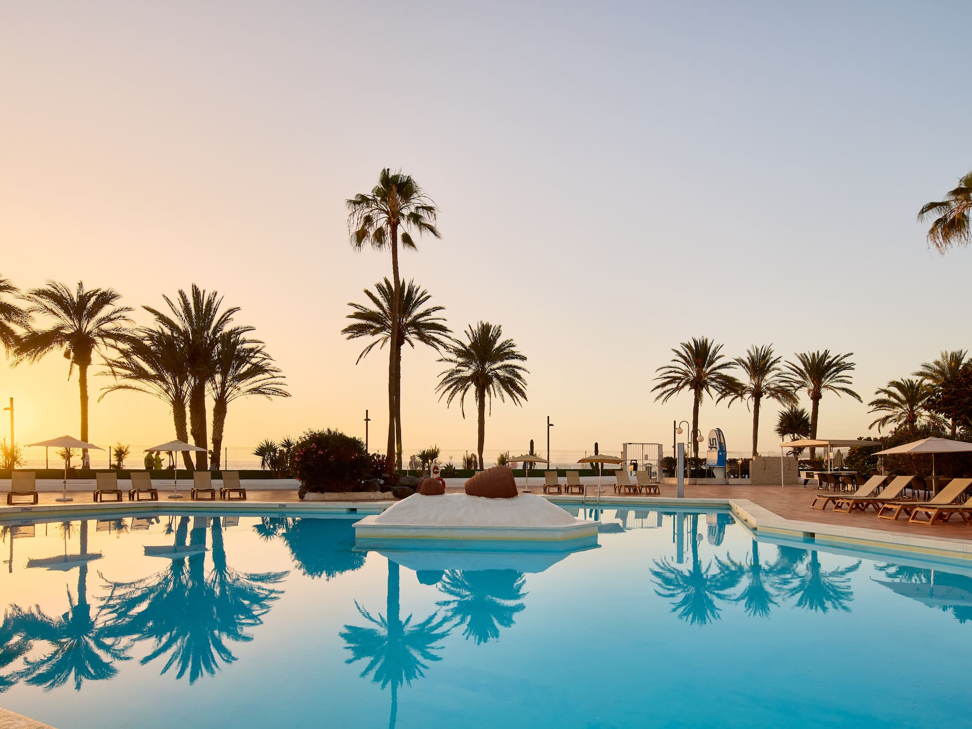 a pool with palm trees and a beach and a sunset