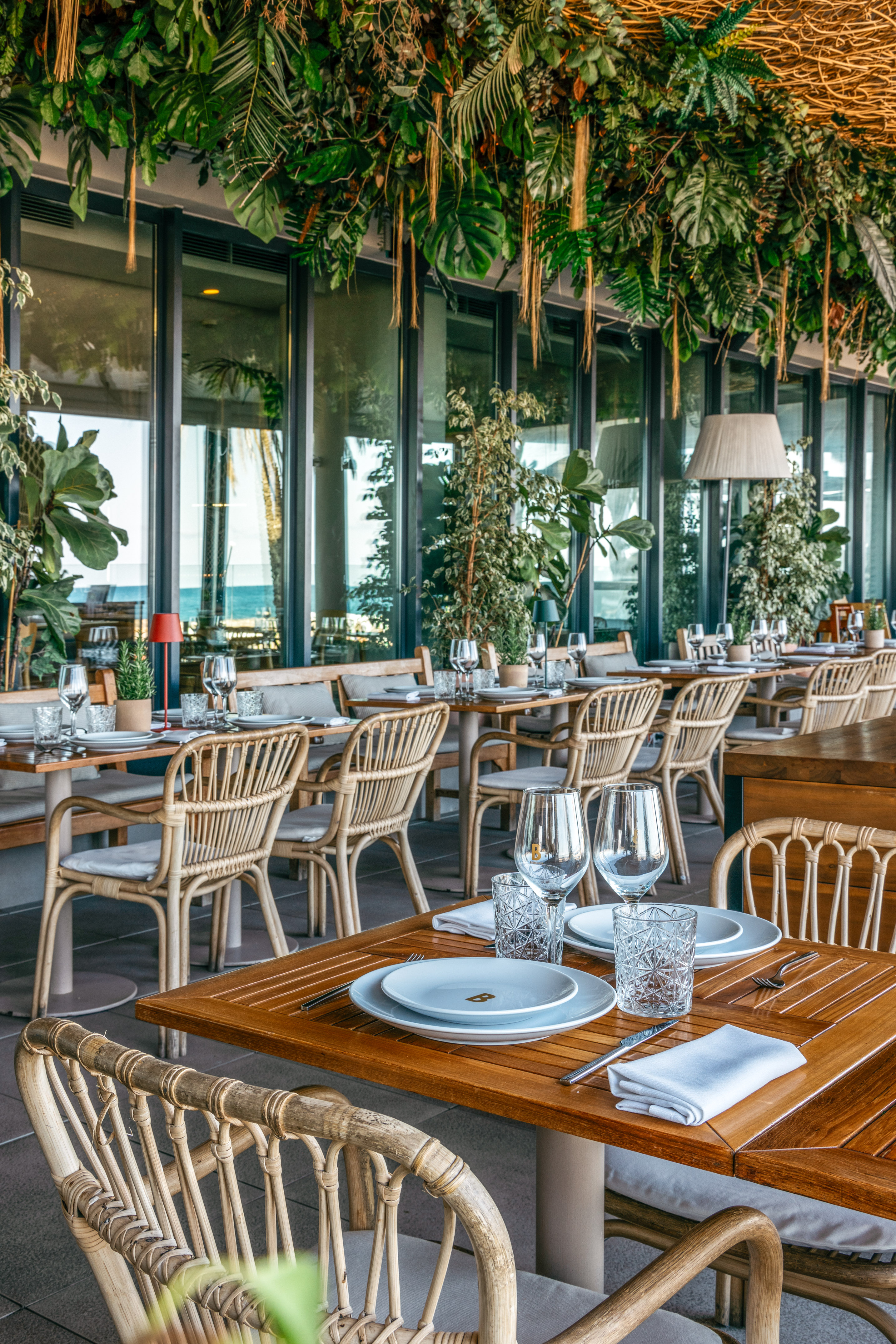 Outdoor restaurant patio with rattan furniture, tropical plants, and ocean view.