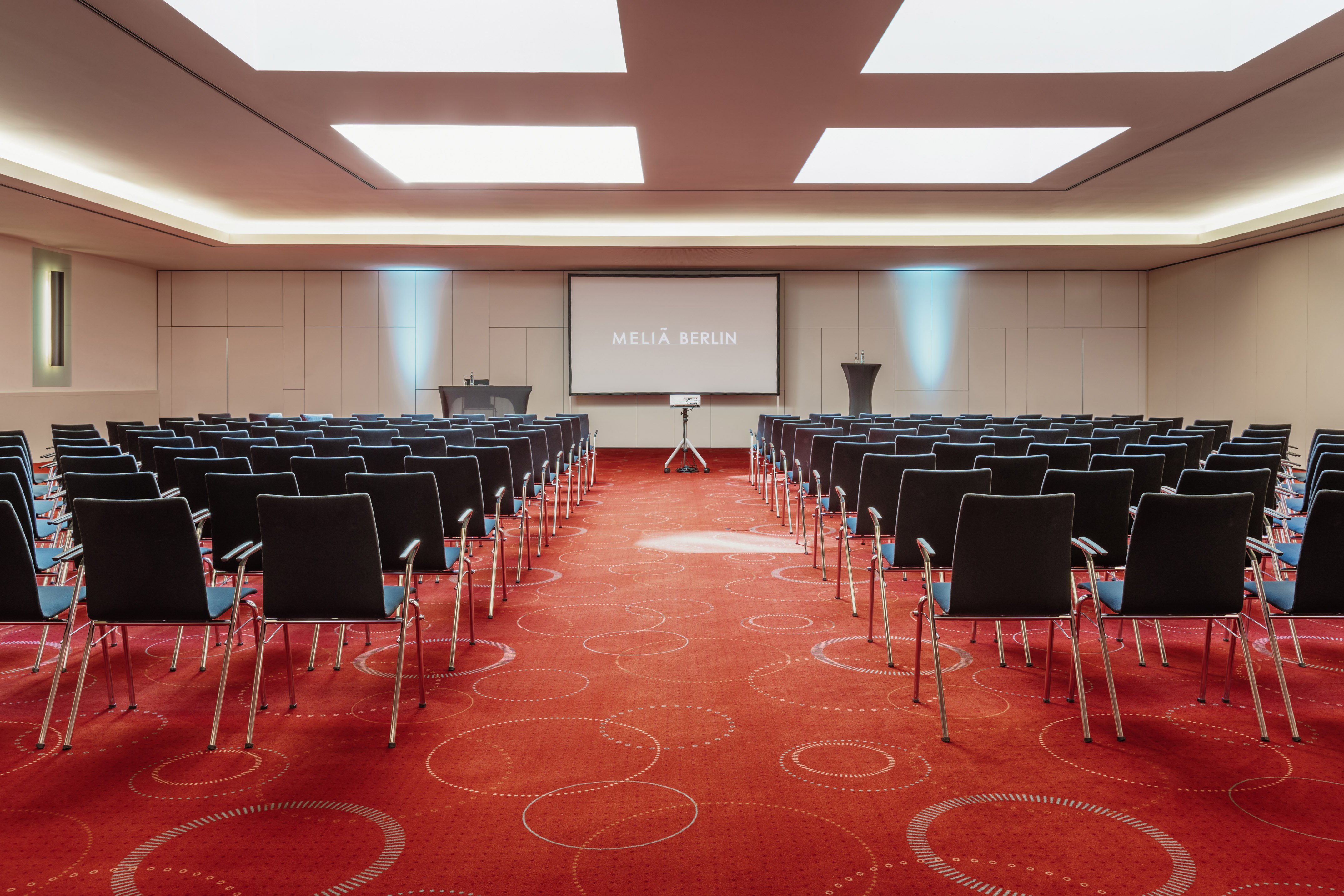 Empty Meliá Berlin conference room with screen, projector, and rows of chairs.