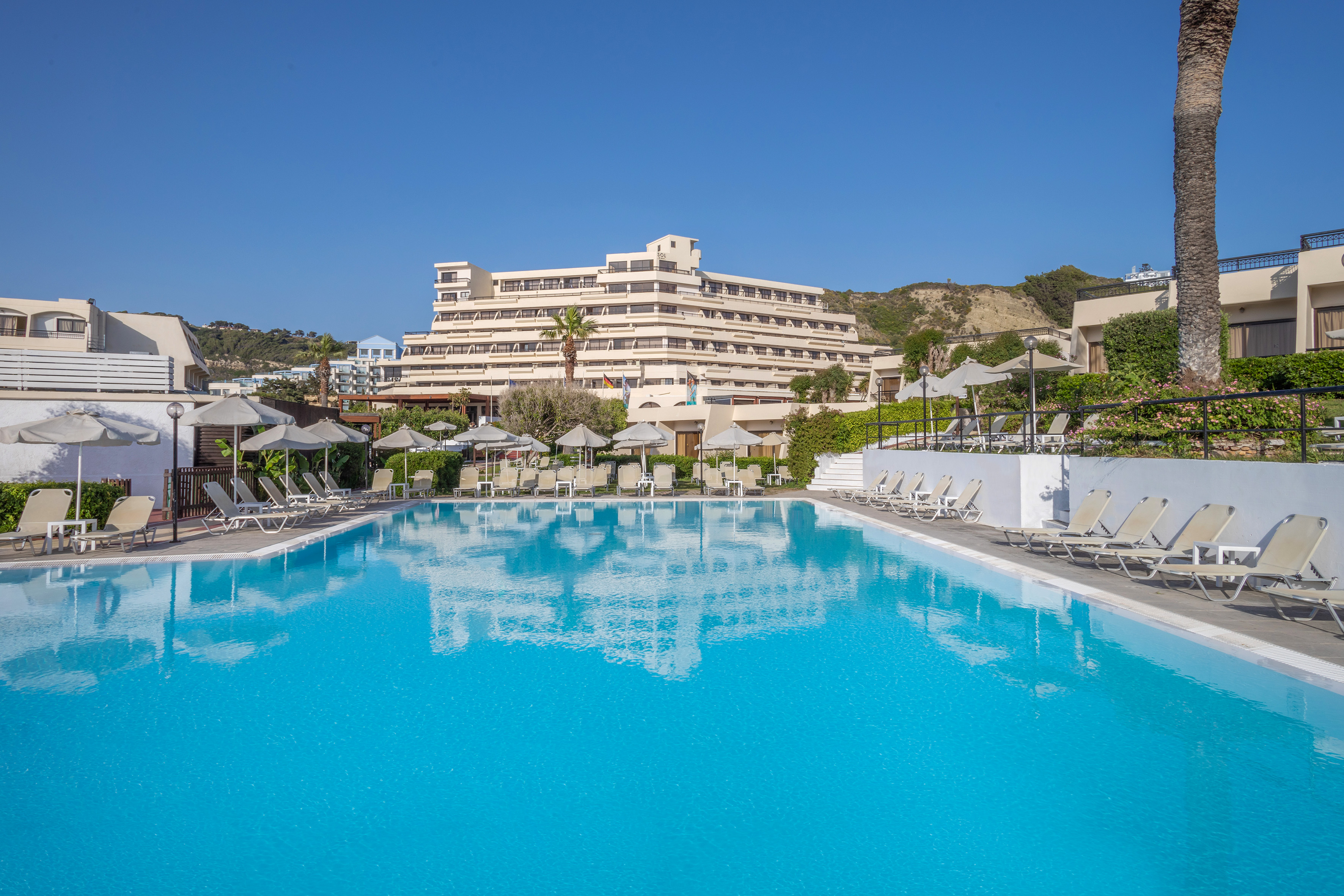 Sunny resort swimming pool with hotel, sun loungers, and palm trees.
