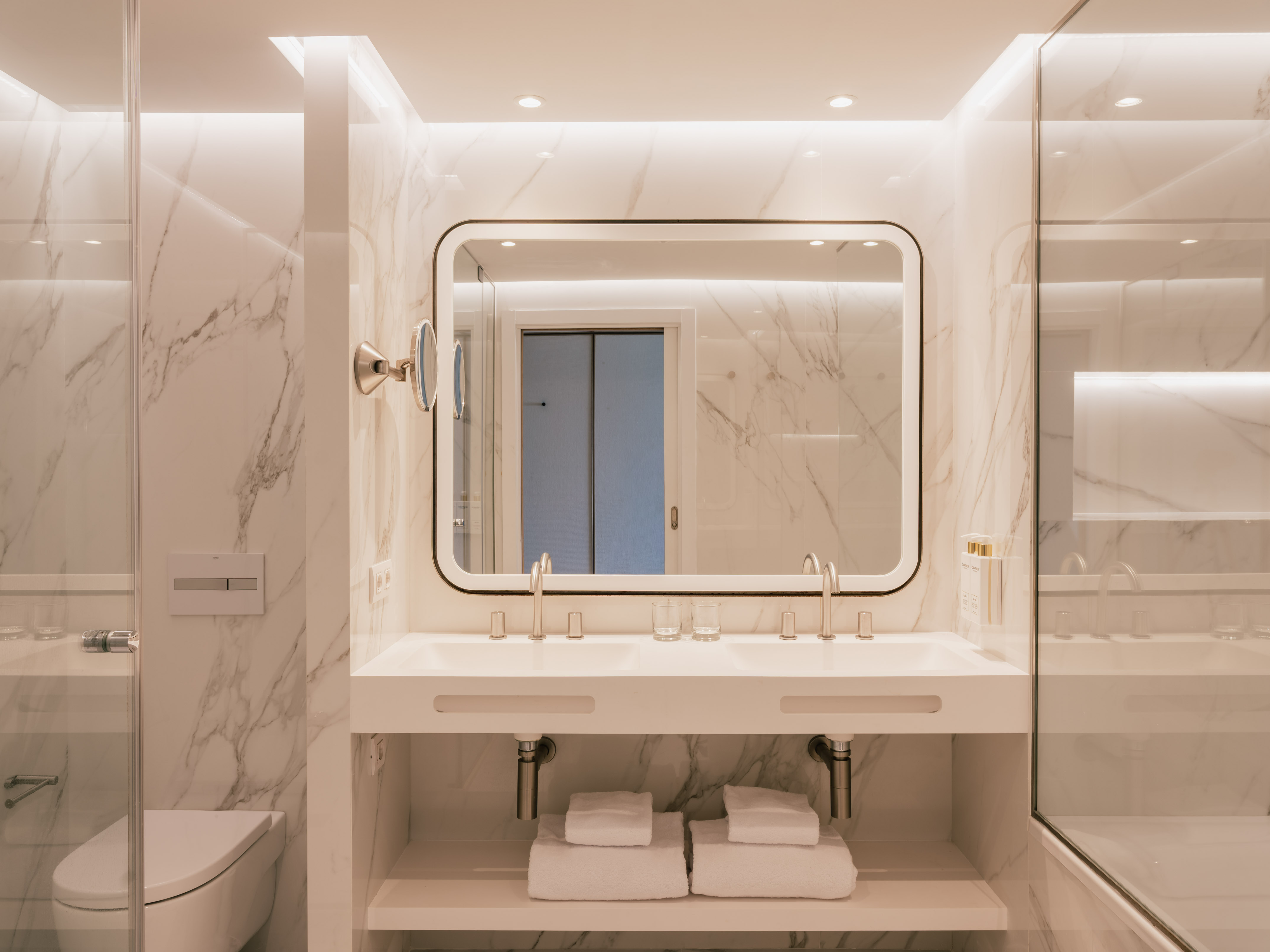 Baño moderno de mármol blanco con doble lavabo y espejo iluminado.