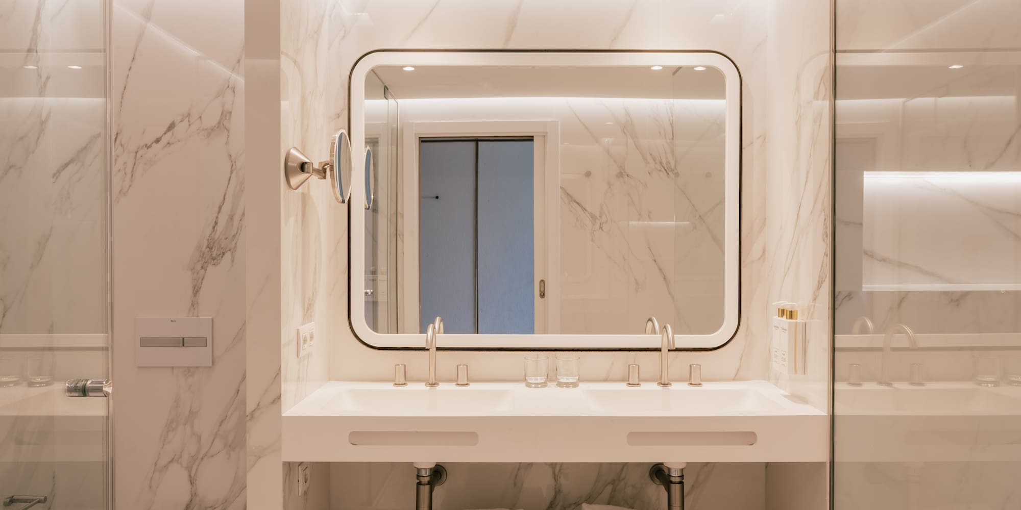 Modern bathroom with white marble walls, double vanity, and backlit mirror.