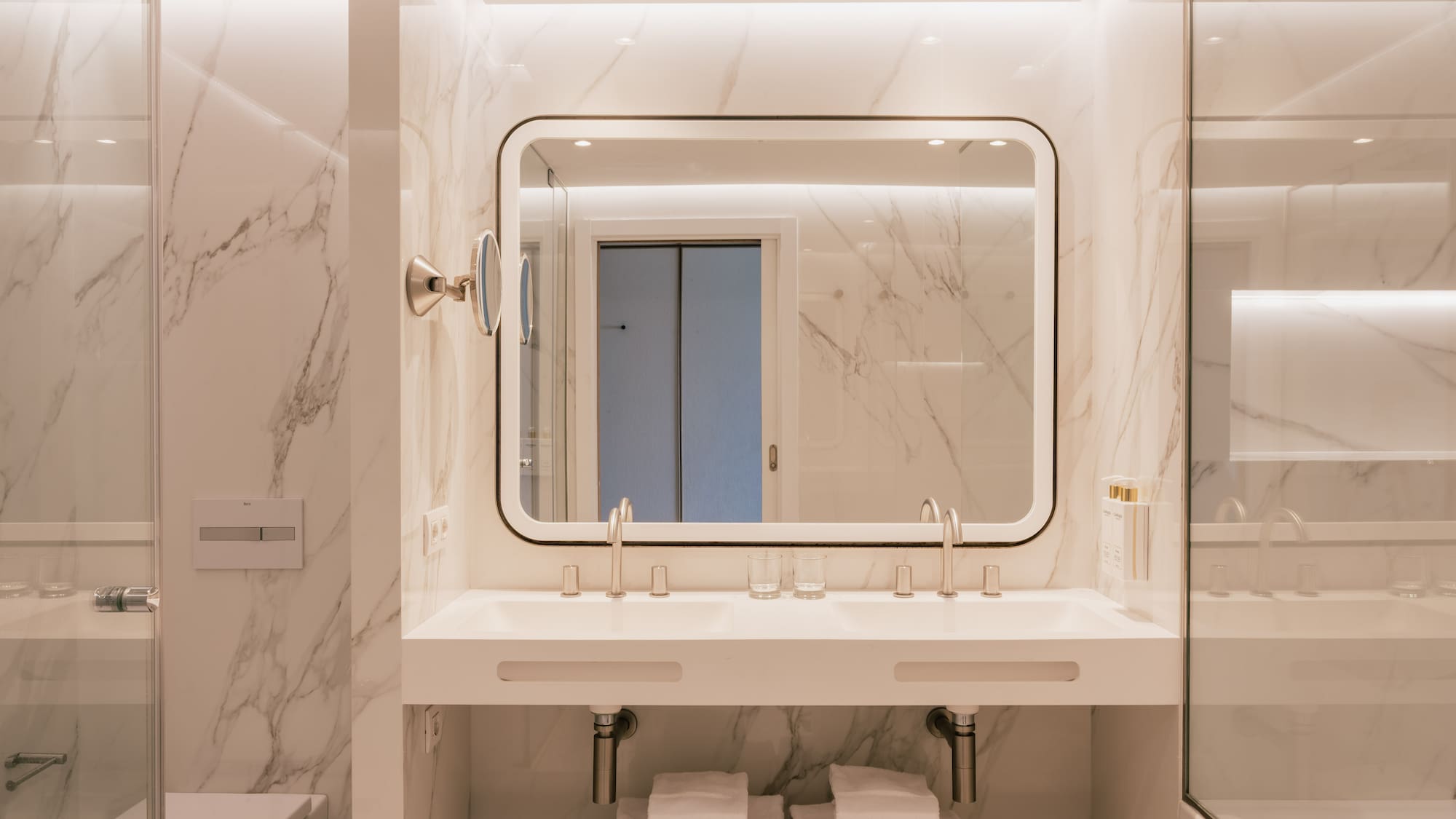Modern bathroom with white marble walls, double vanity, and backlit mirror.