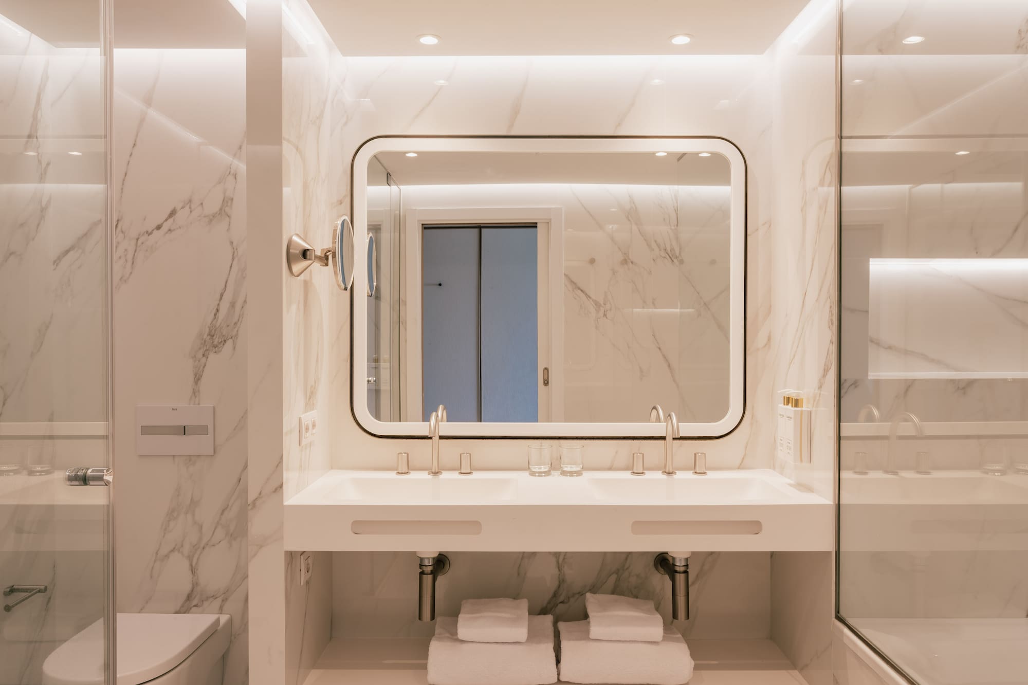 Baño moderno de mármol blanco con doble lavabo y espejo iluminado.
