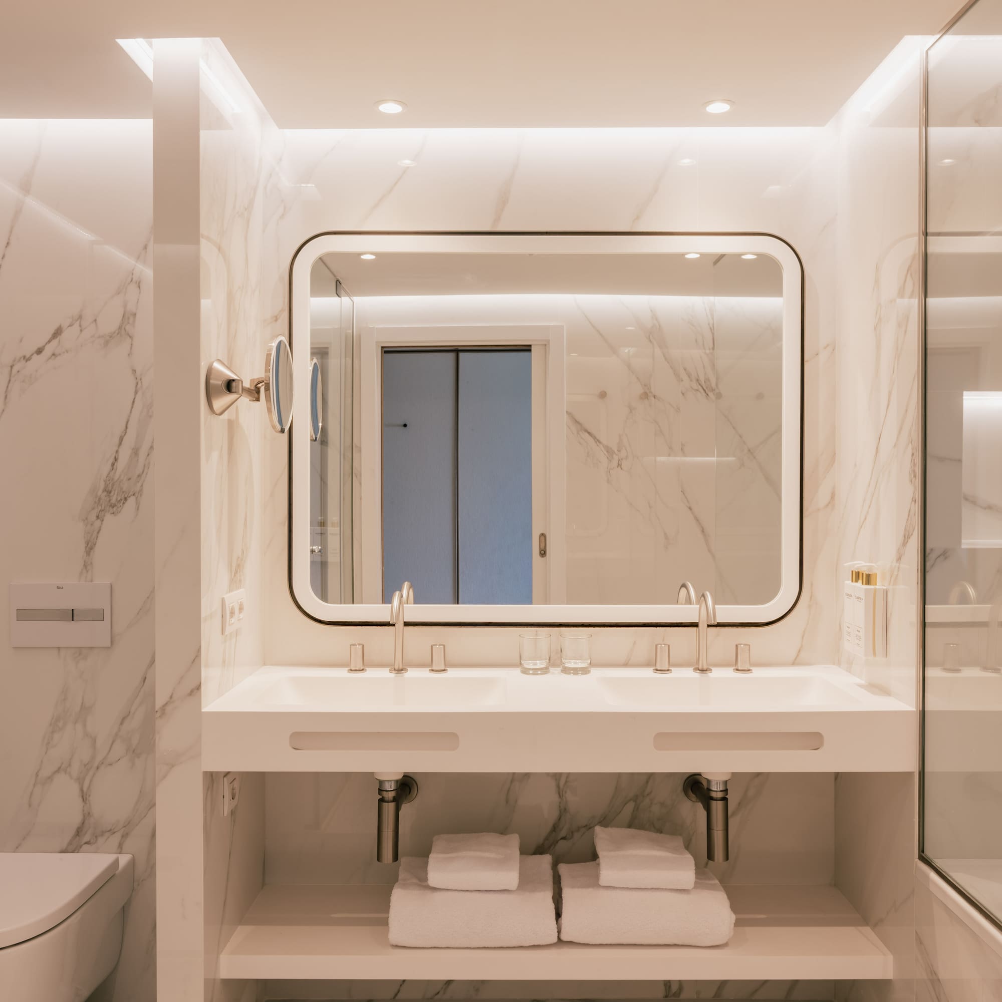 Modern bathroom with white marble walls, double vanity, and backlit mirror.