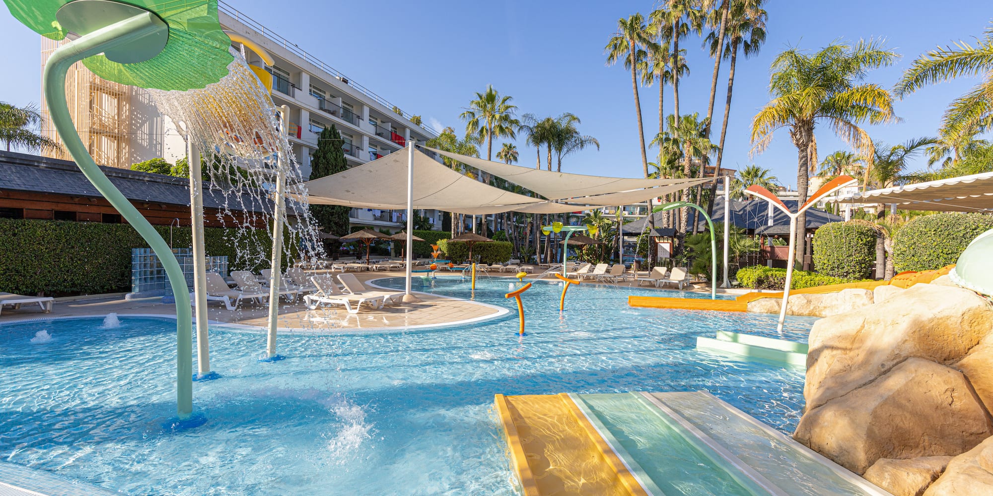Sunny hotel pool with water slides, splash pad, and palm trees.