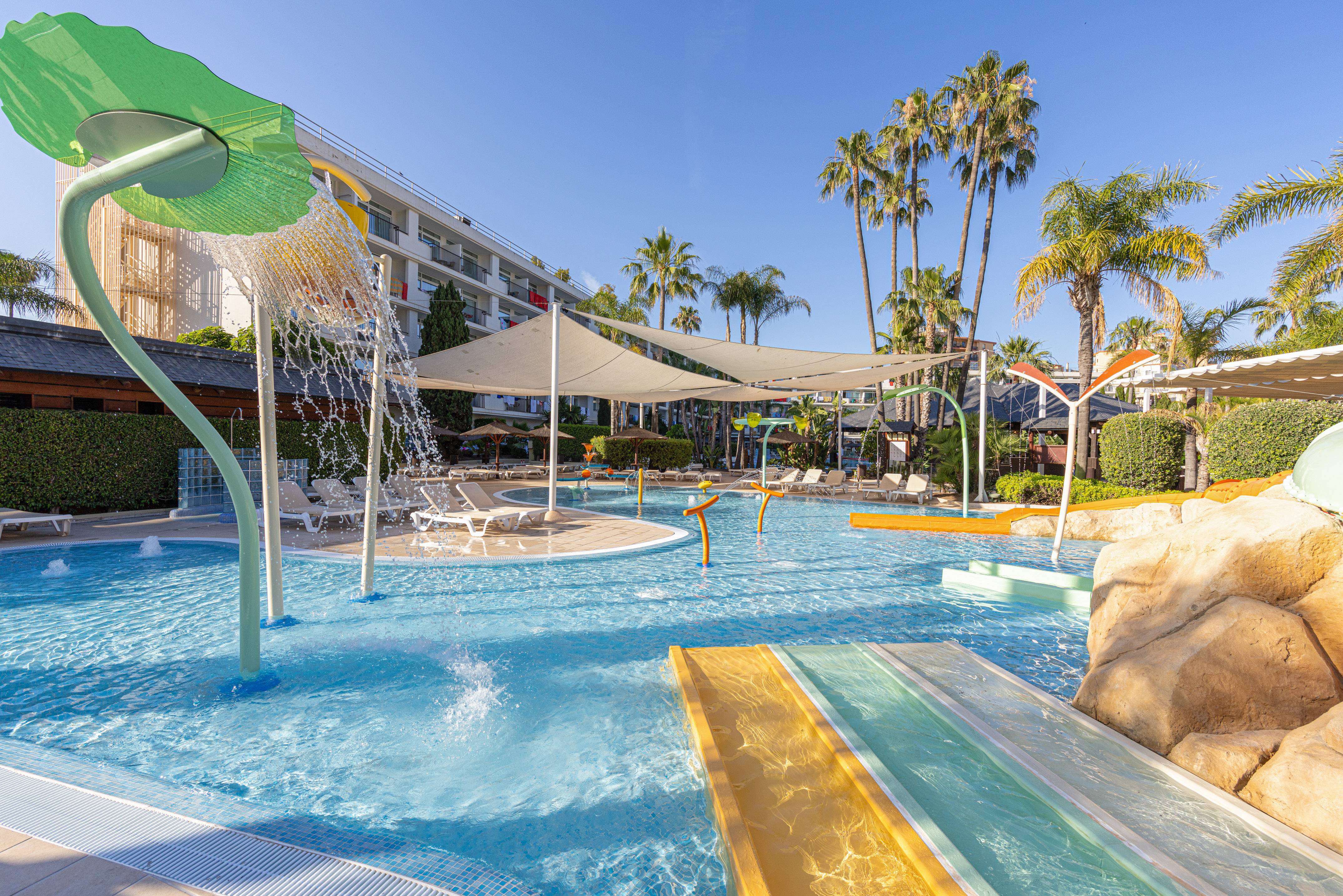 Sunny hotel pool with water slides, splash pad, and palm trees.