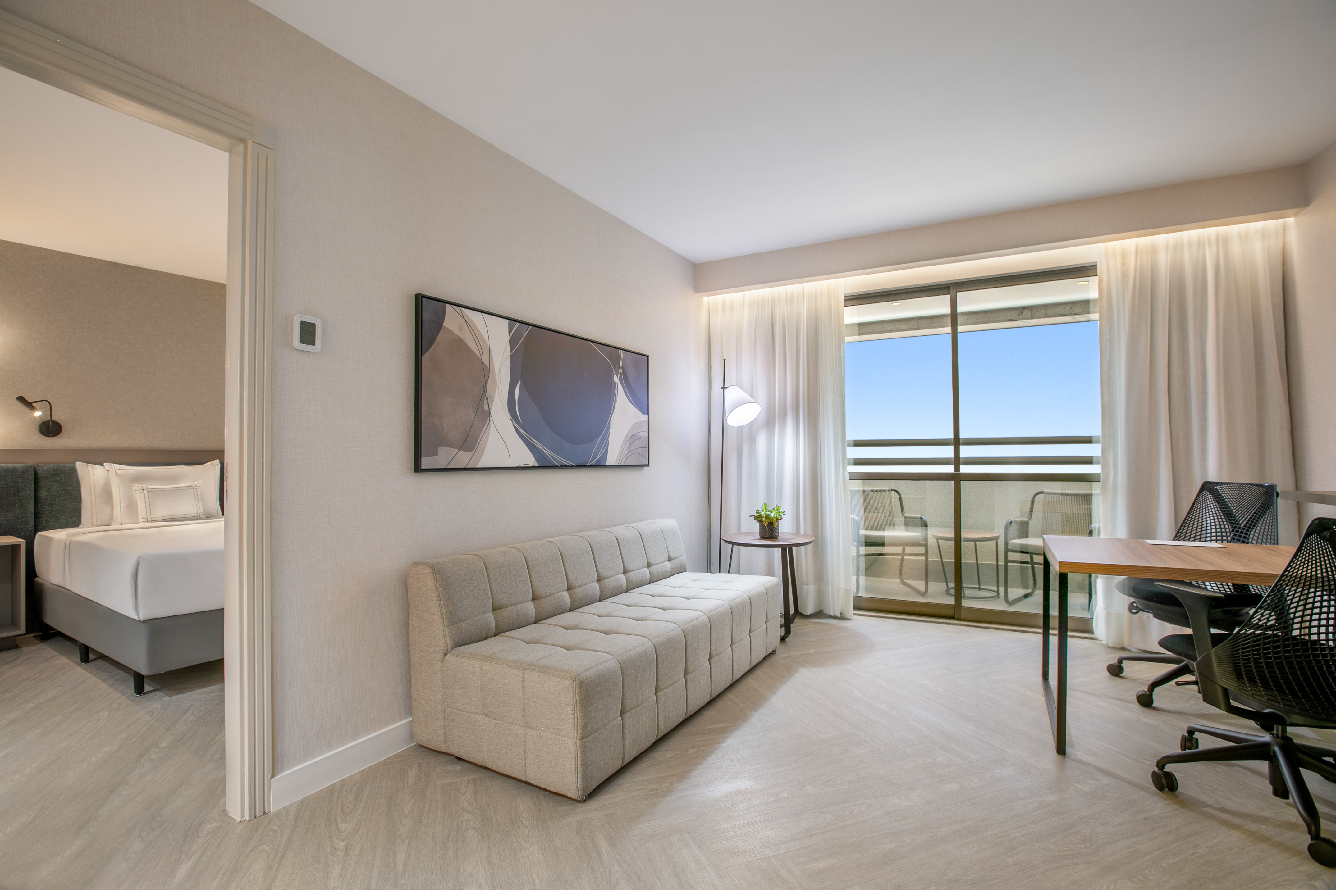 Modern hotel suite with living area, desk workstation, balcony view, and adjoining bedroom.