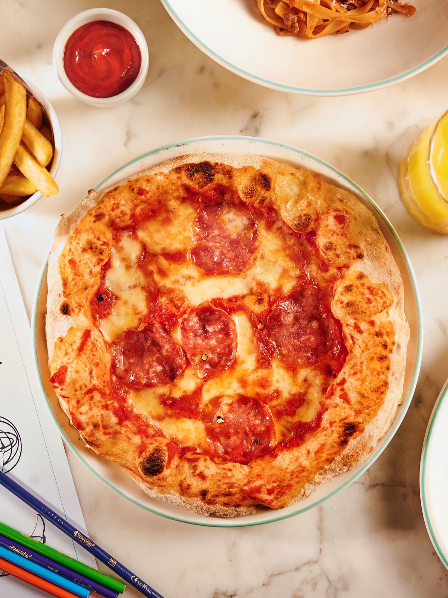 Family dining with salami pizza, fries, pasta, juice, and activity sheet.