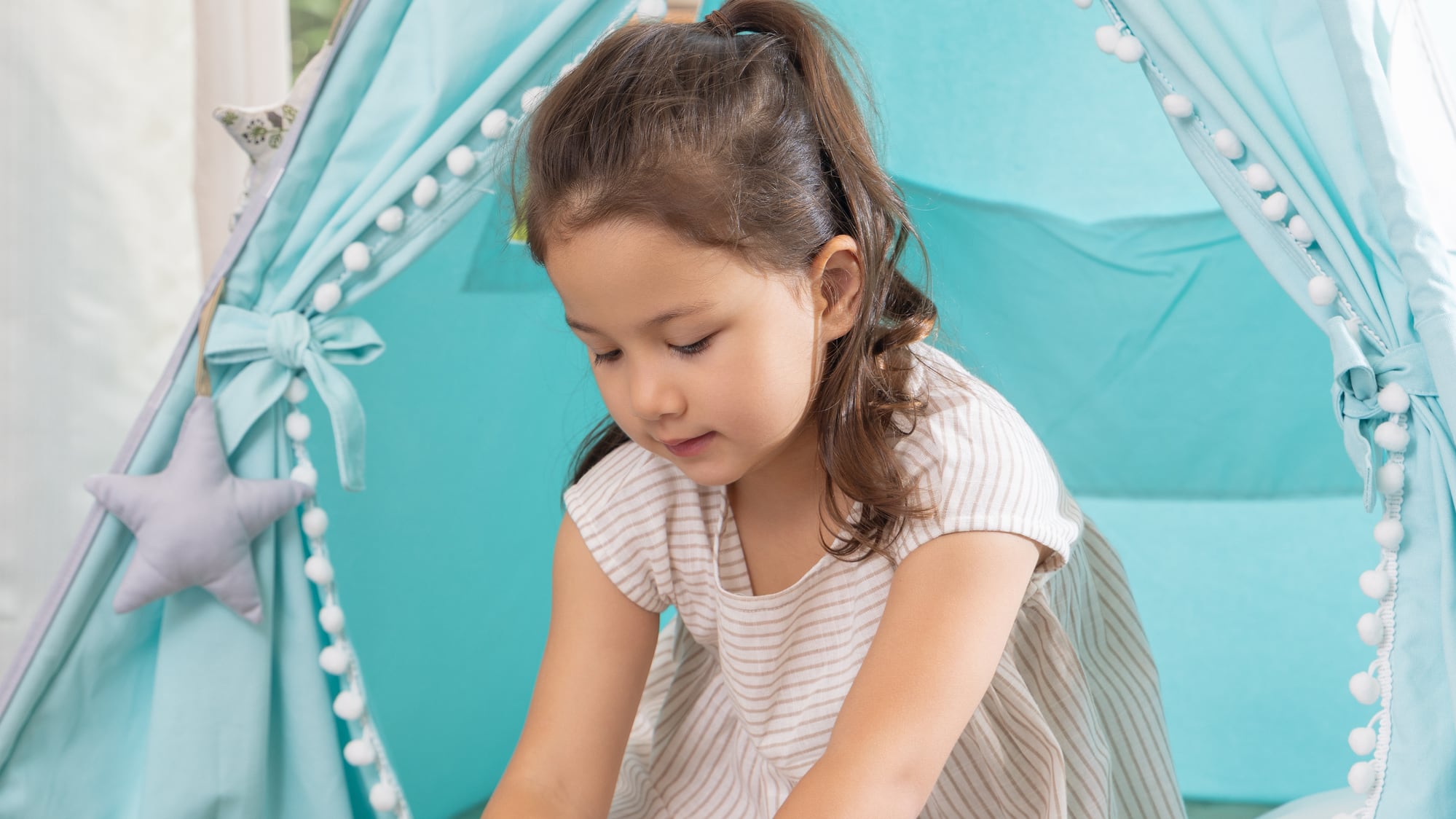 Young girl playing a puzzle inside a blue teepee tent with pom-pom trim.