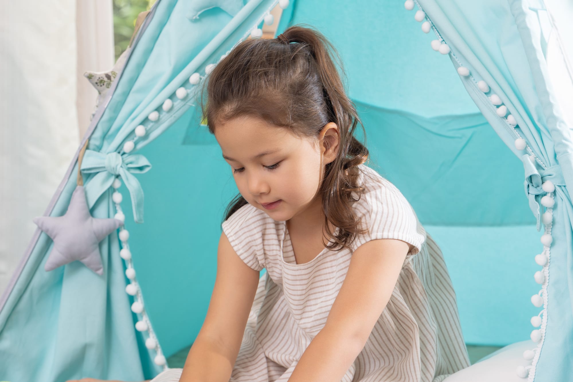 Young girl playing a puzzle inside a blue teepee tent with pom-pom trim.