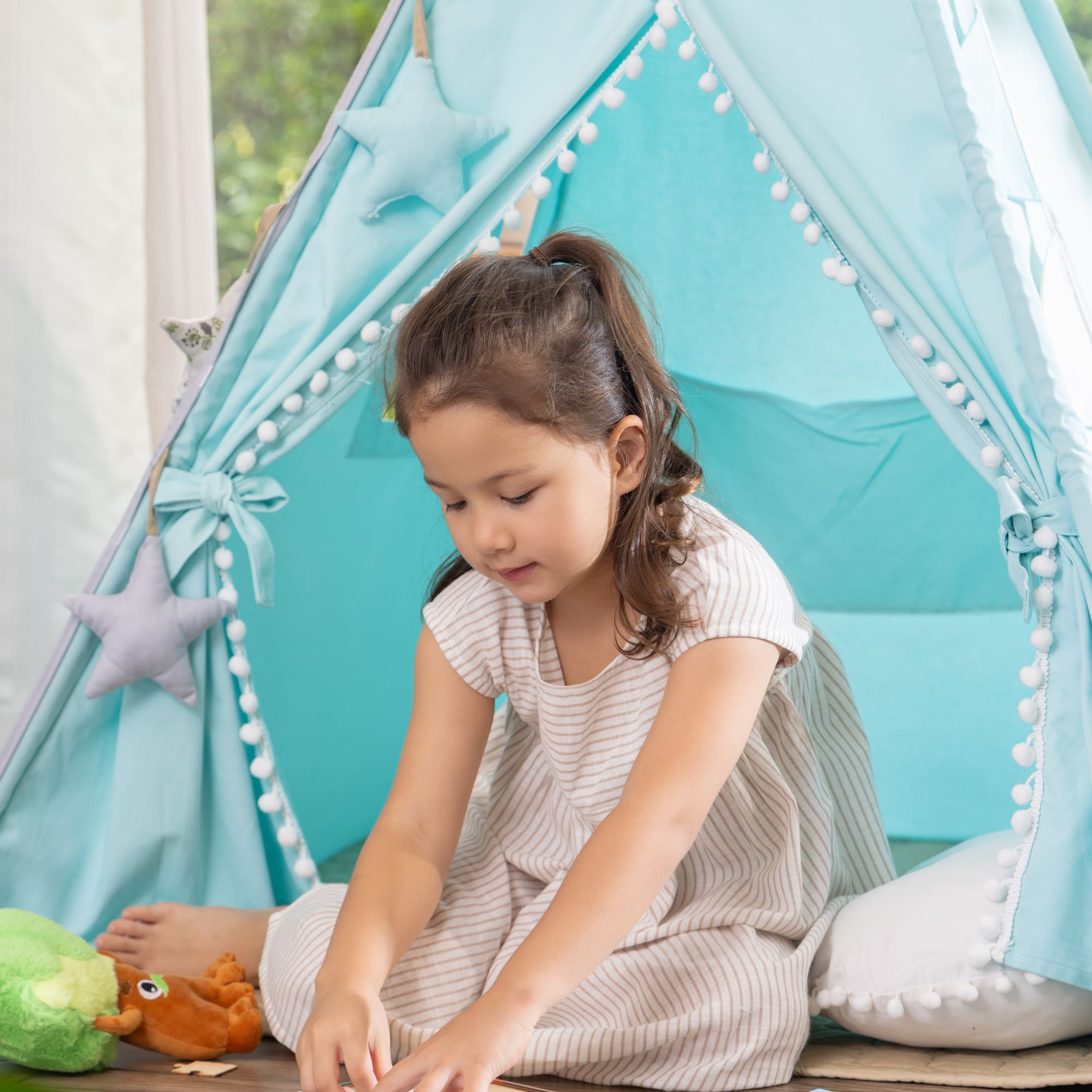 Young girl playing a puzzle inside a blue teepee tent with pom-pom trim.