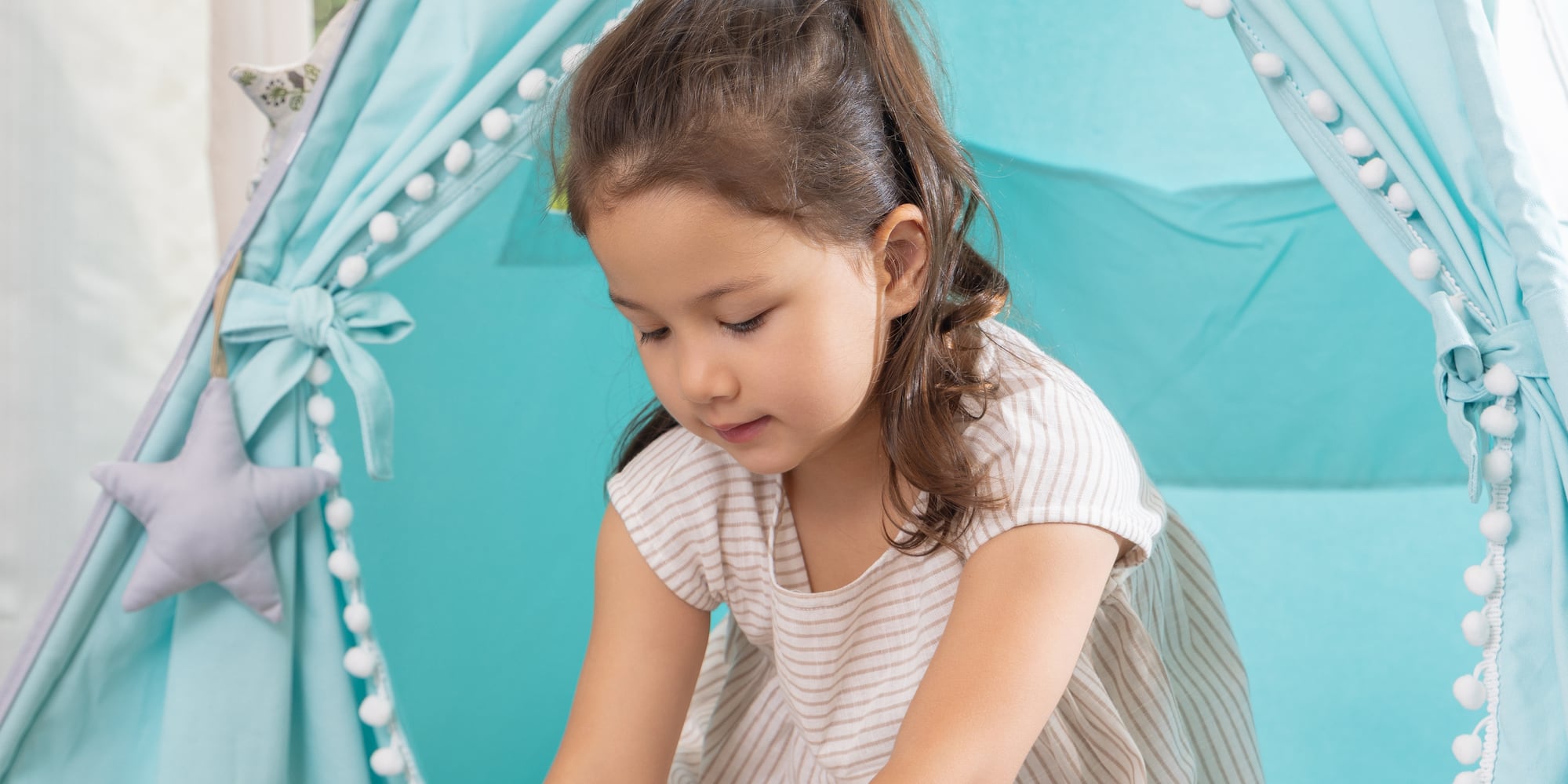 Young girl playing a puzzle inside a blue teepee tent with pom-pom trim.