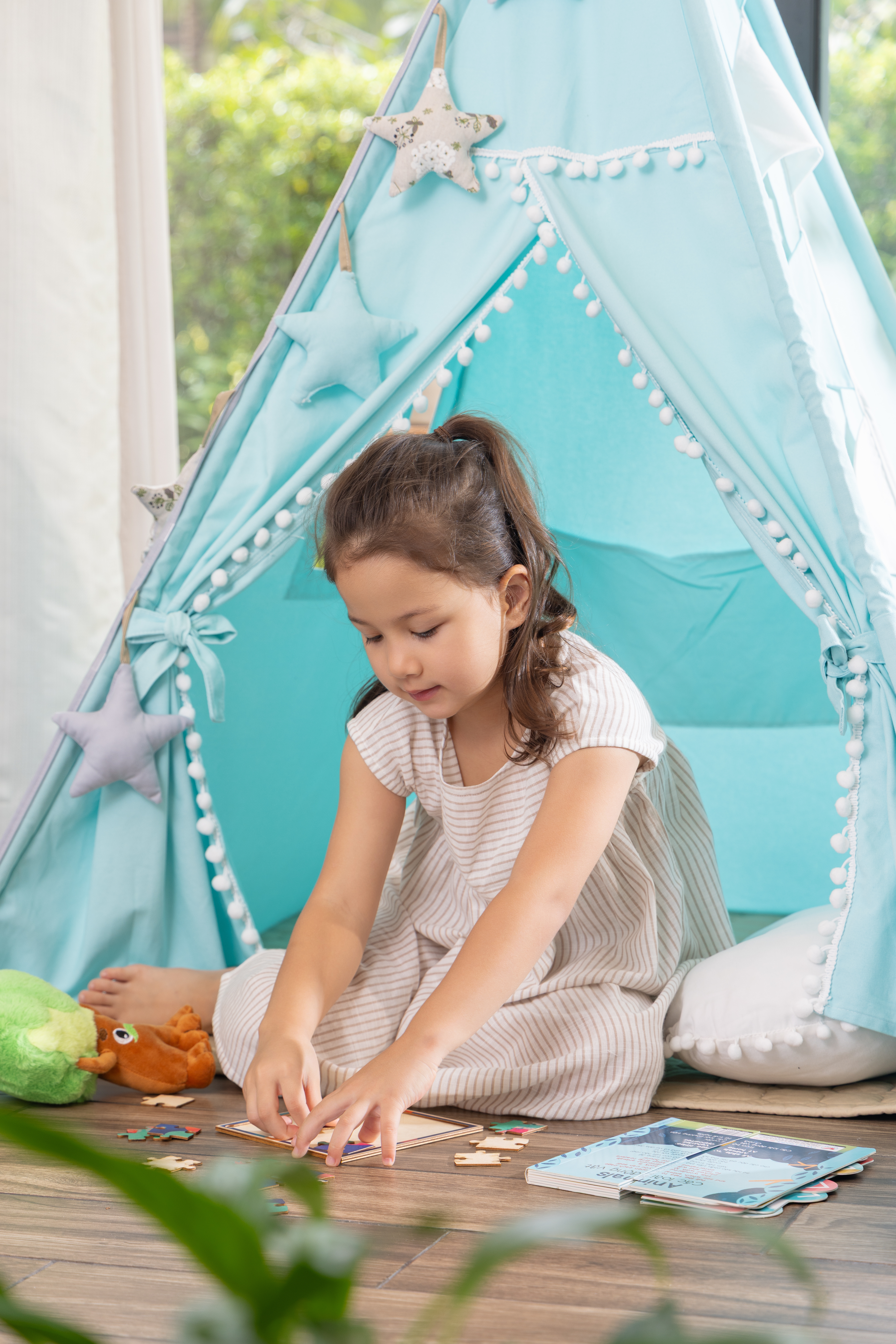 Young girl playing a puzzle inside a blue teepee tent with pom-pom trim.