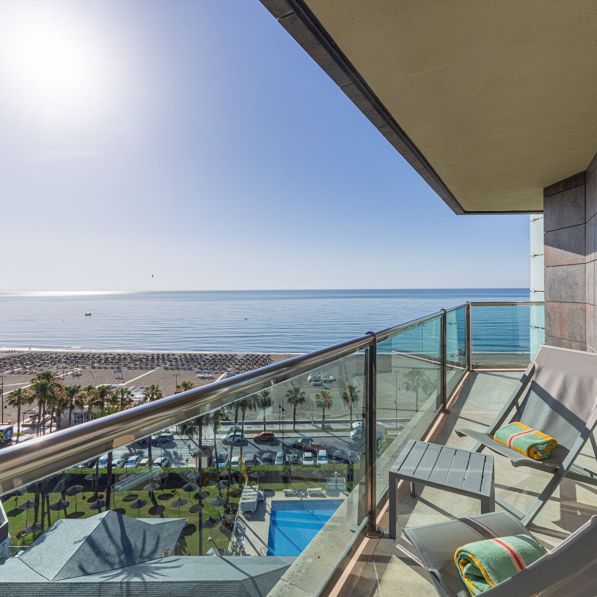 Hotel balcony with chairs overlooking sunny beach, sea, palm trees, and pool.