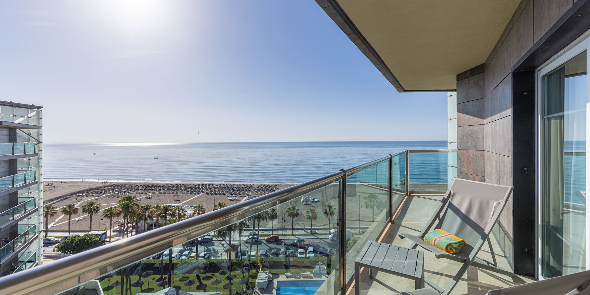 Hotel balcony with chairs overlooking sunny beach, sea, palm trees, and pool.