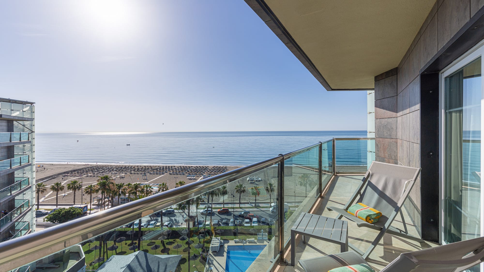 Sunny hotel balcony with sea view, beach, palm trees, and swimming pool.