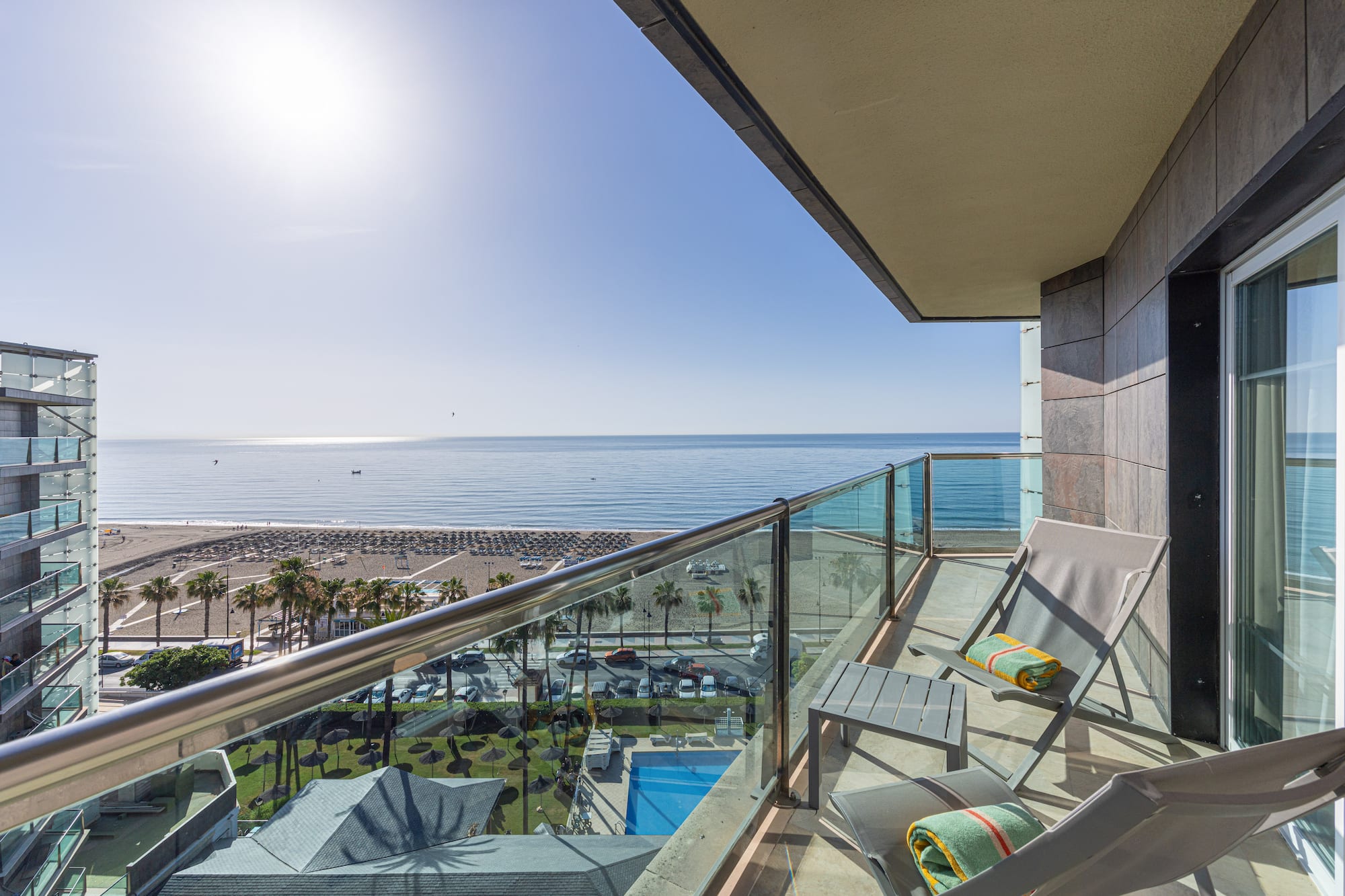 Modern balcony with ocean view, beach, palm trees, and resort pool.