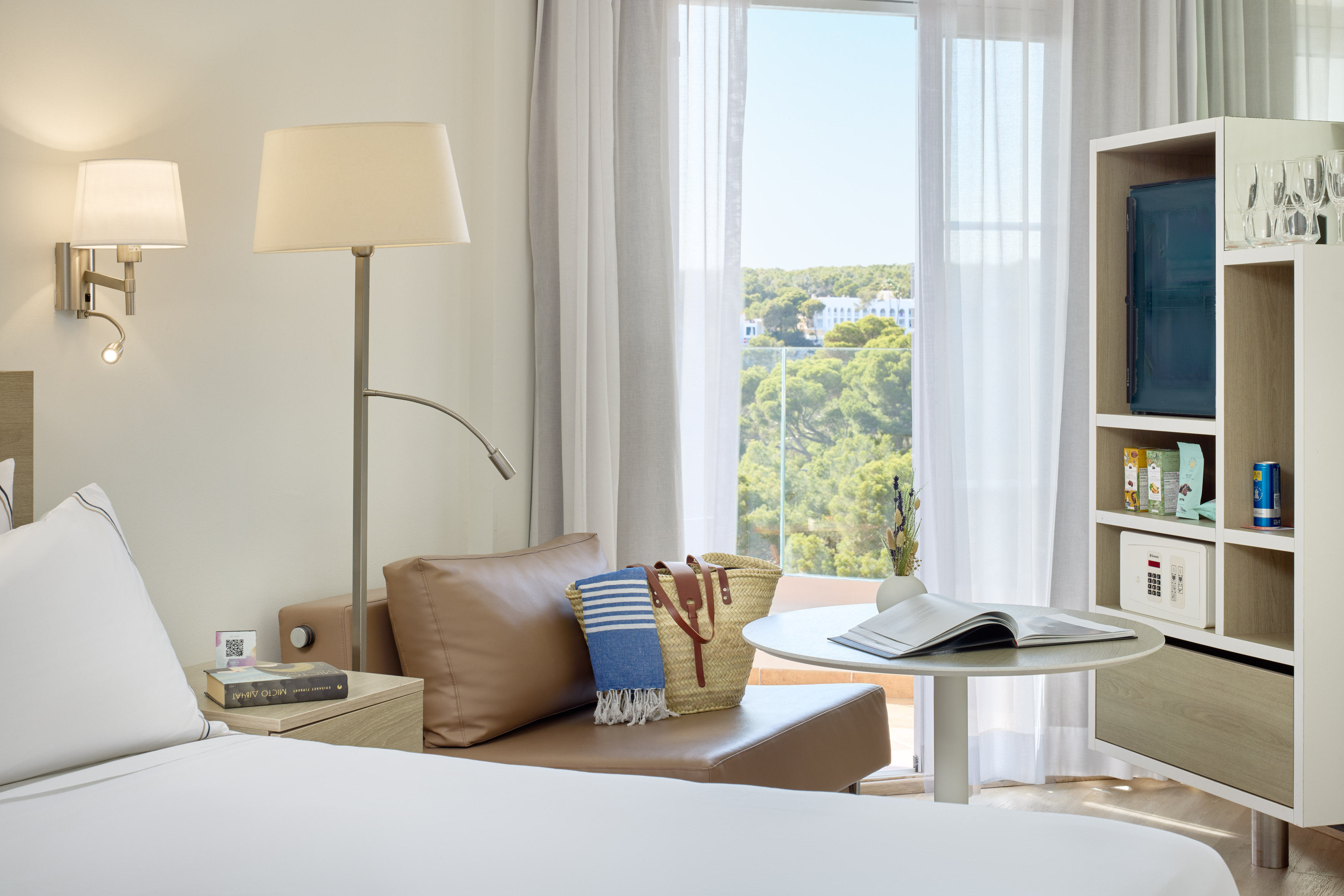 Modern hotel room with balcony, lush green view, and comfortable daybed.