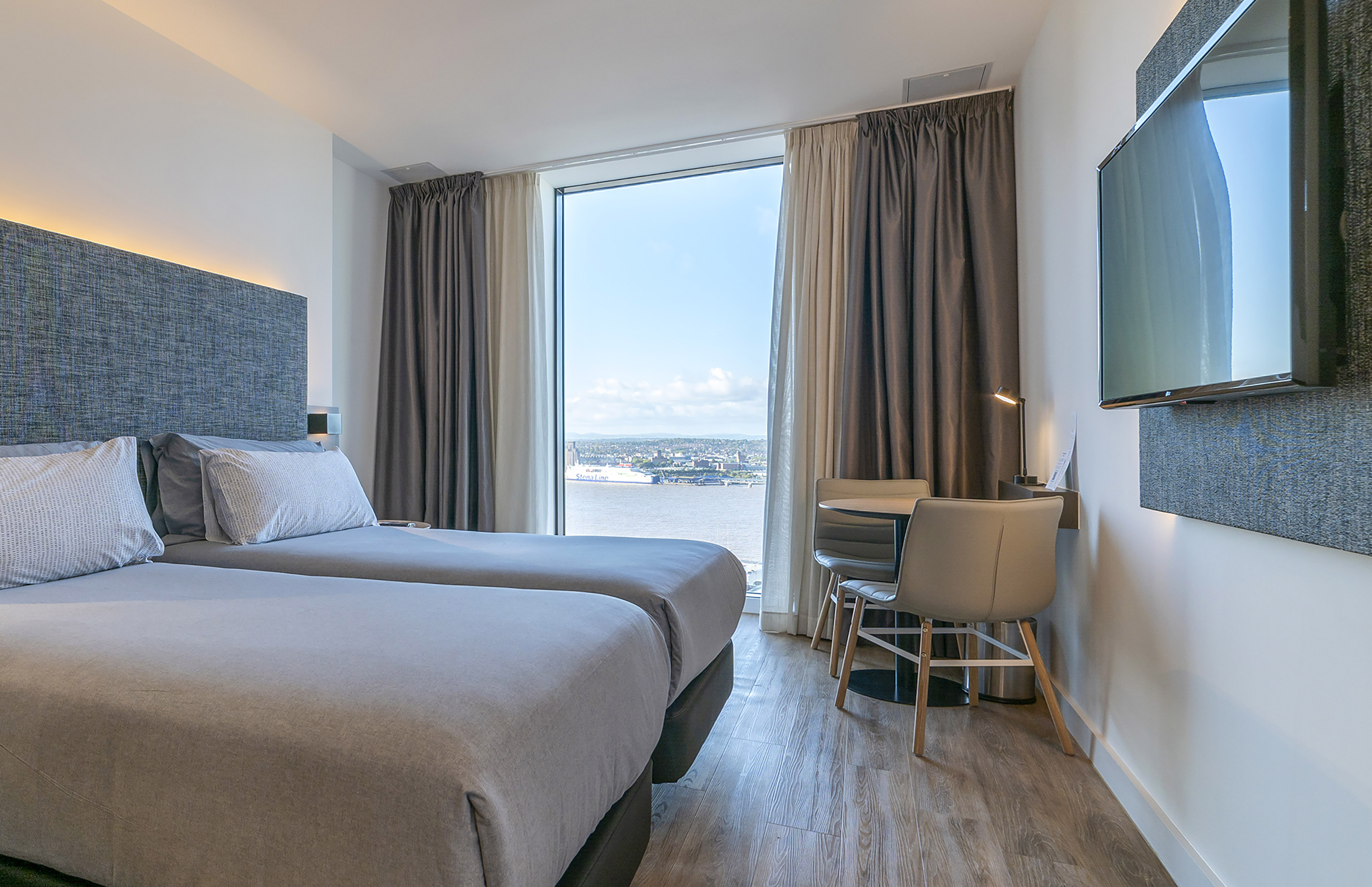 Modern twin hotel room with city and river view from large window.