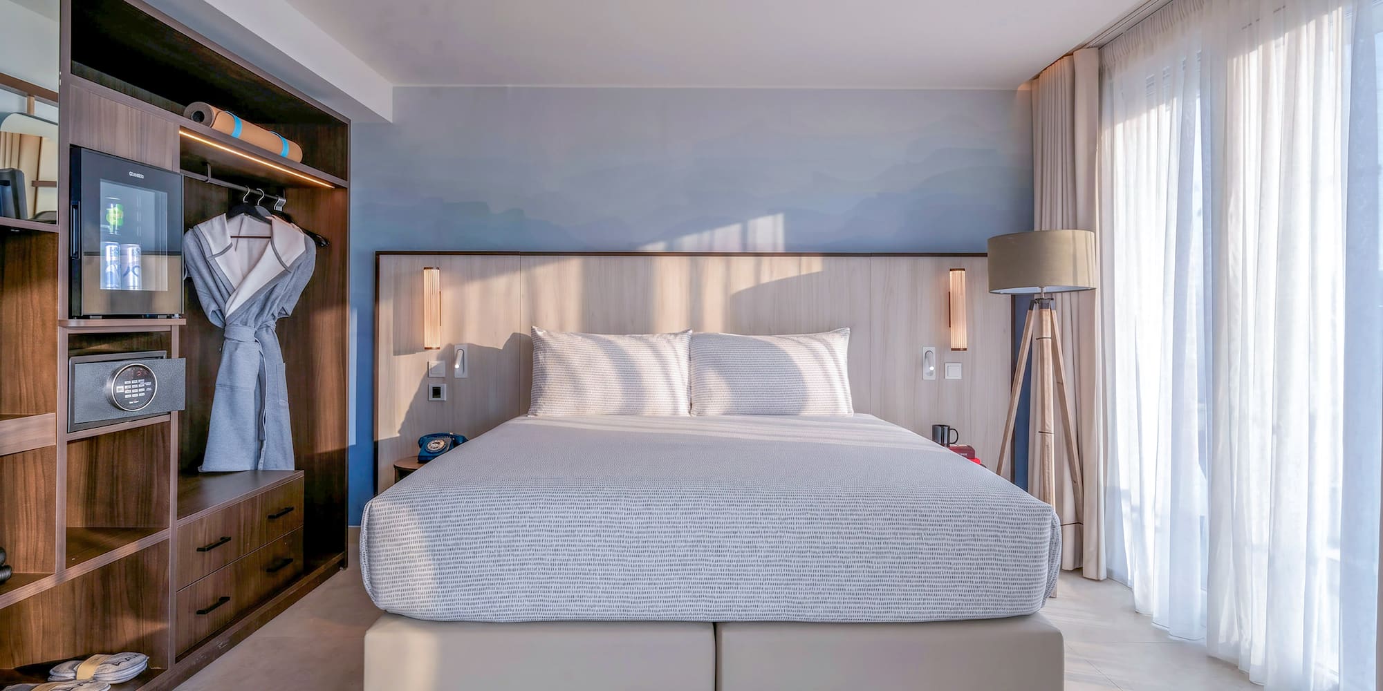 Modern hotel bedroom: bed, built-in wooden wardrobe, mini-bar, and bright natural light.