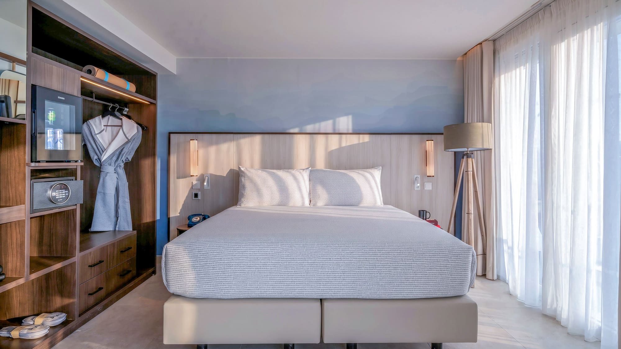 Modern hotel bedroom: bed, built-in wooden wardrobe, mini-bar, and bright natural light.
