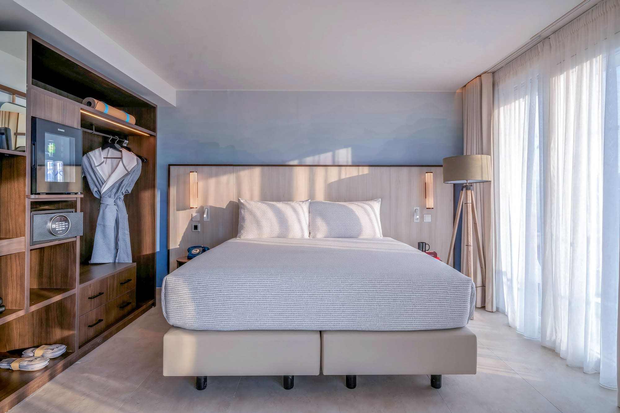 Modern hotel bedroom: bed, built-in wooden wardrobe, mini-bar, and bright natural light.