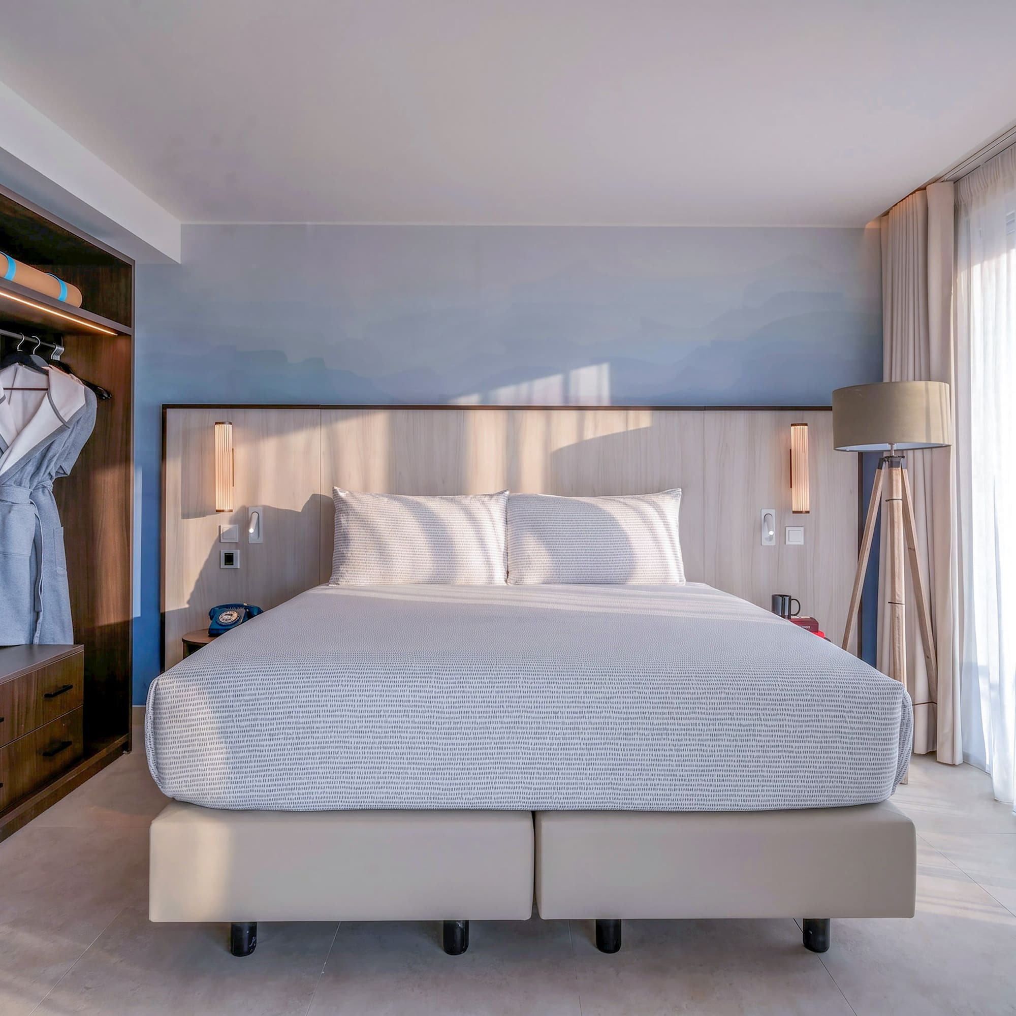 Modern hotel bedroom: bed, built-in wooden wardrobe, mini-bar, and bright natural light.