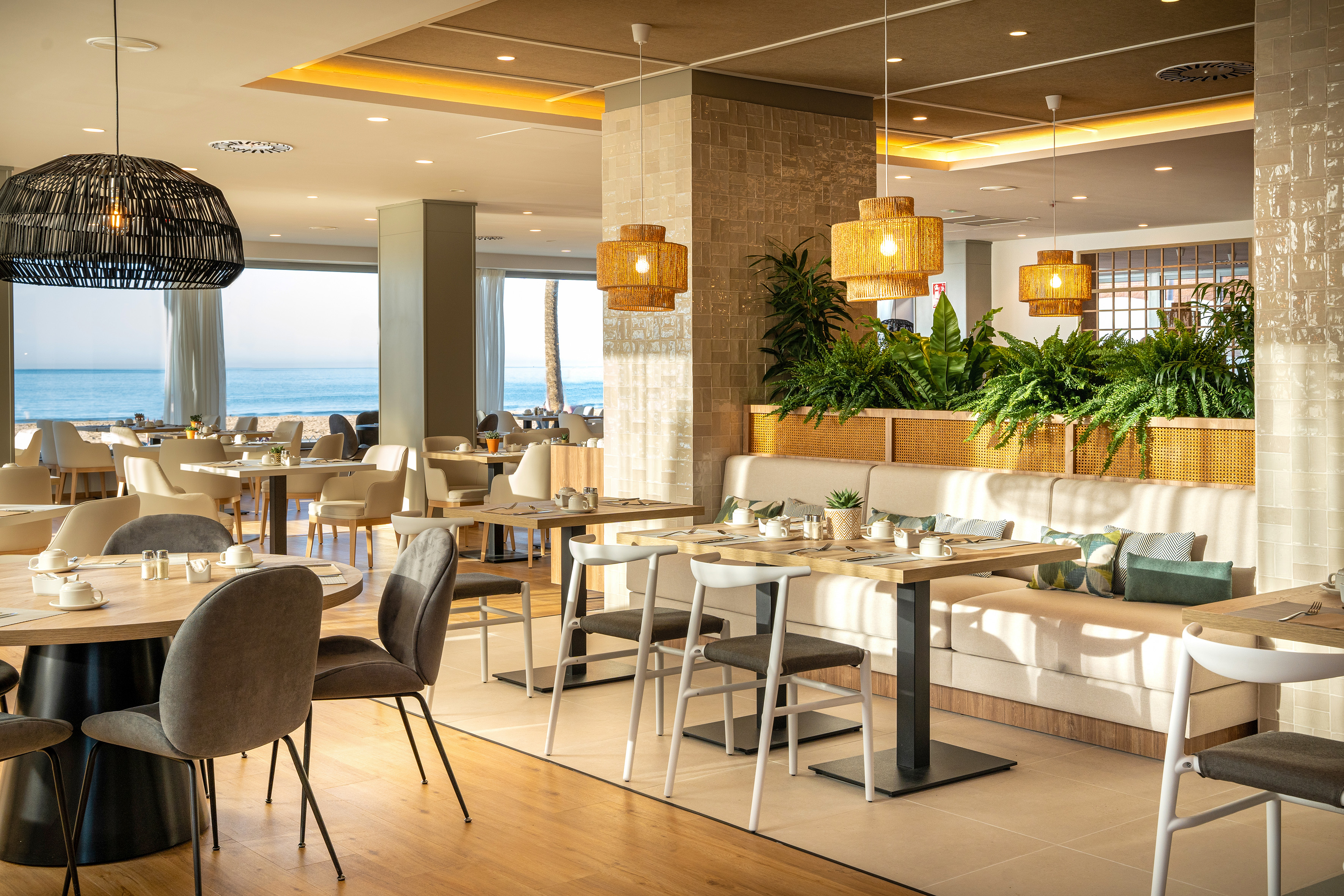 Coastal restaurant with sea view, modern tables, rattan lights, and tropical plants.