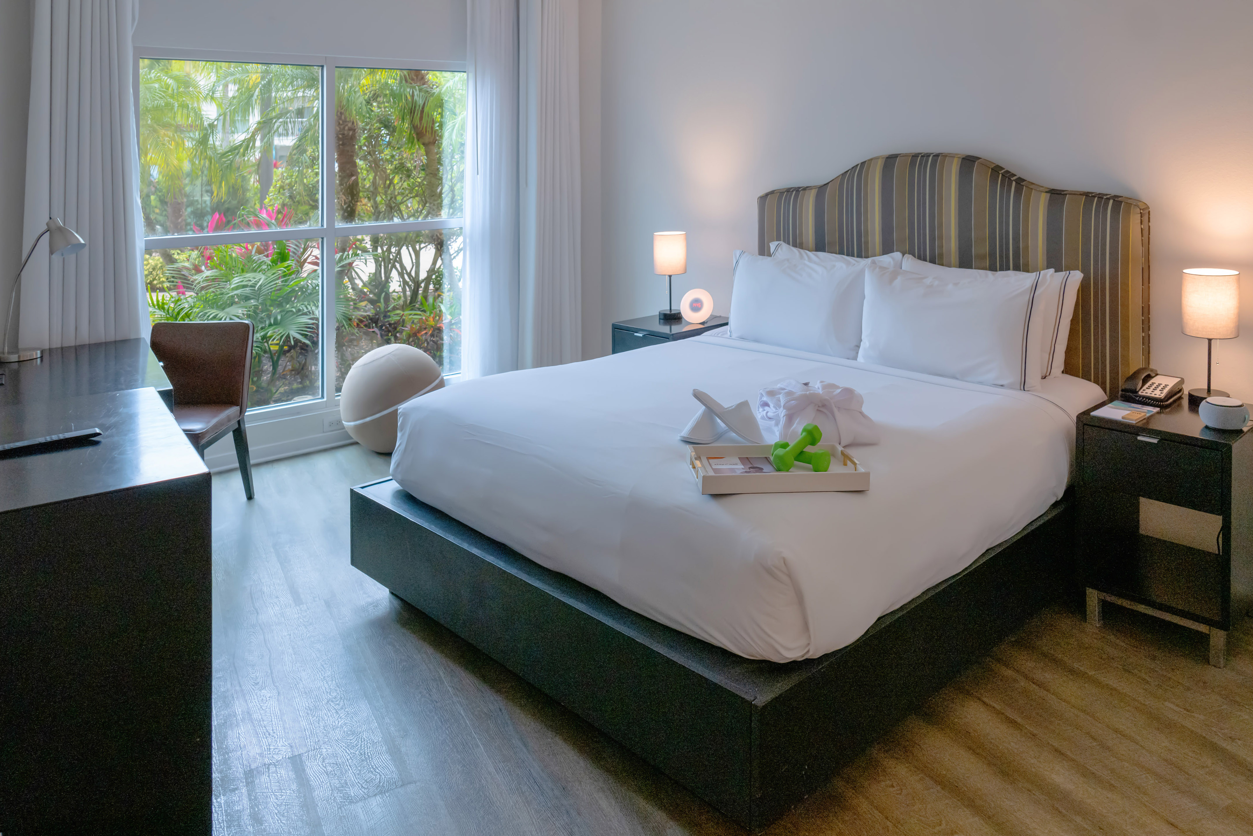 Modern hotel room with a large bed, fitness tray, and window overlooking tropical palms.