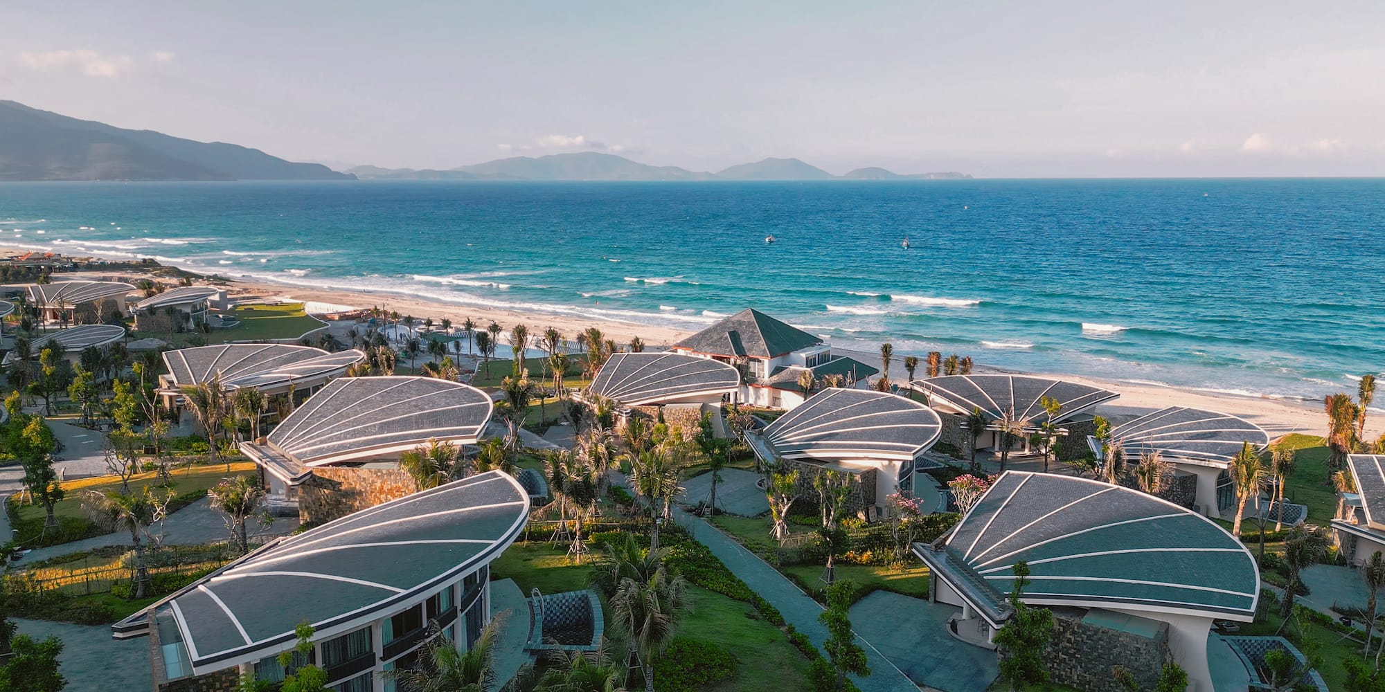 Beachfront resort with unique shell-shaped roofs, turquoise ocean, and mountains.