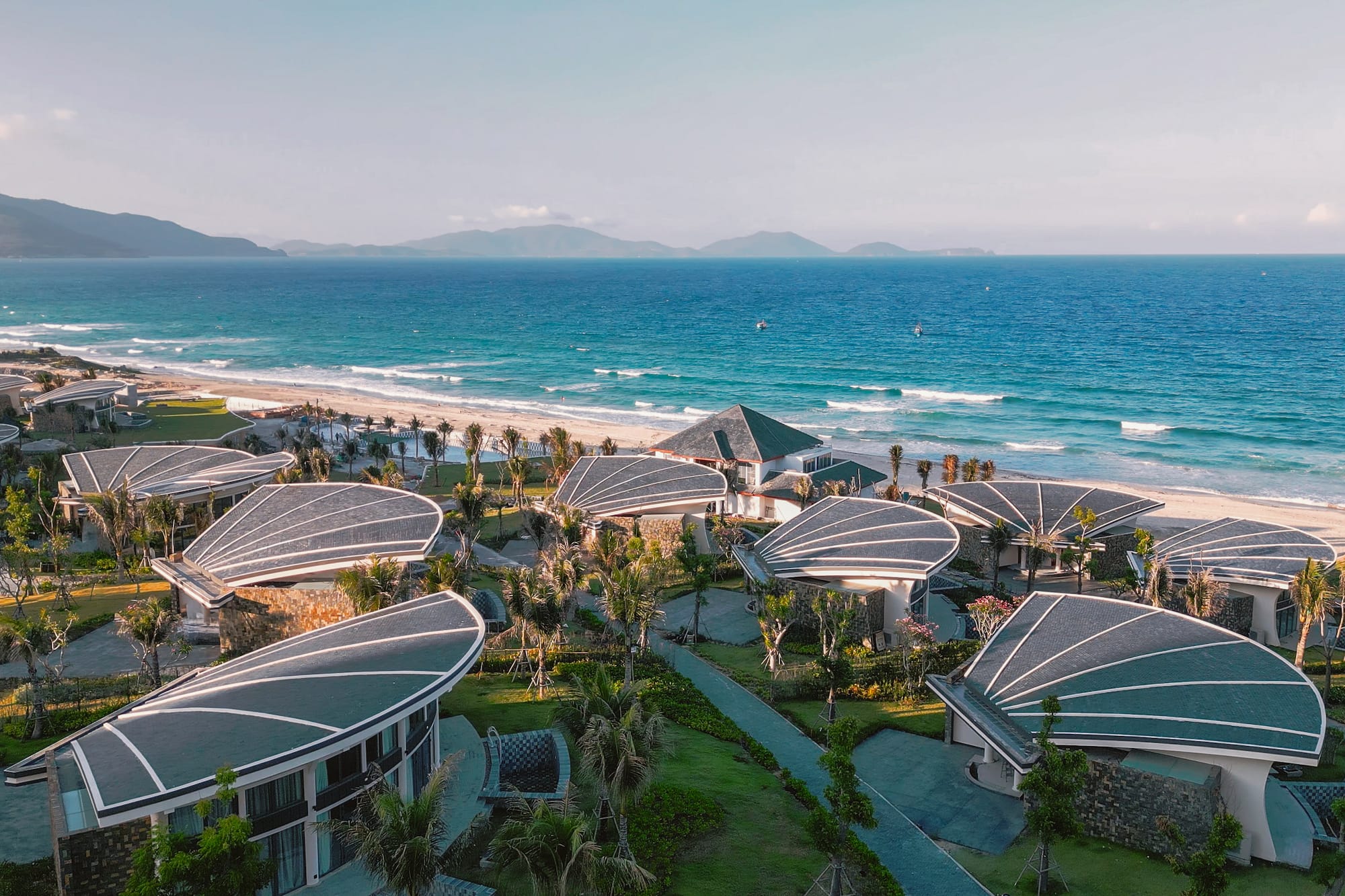 Beachfront resort with unique shell-shaped roofs, turquoise ocean, and mountains.