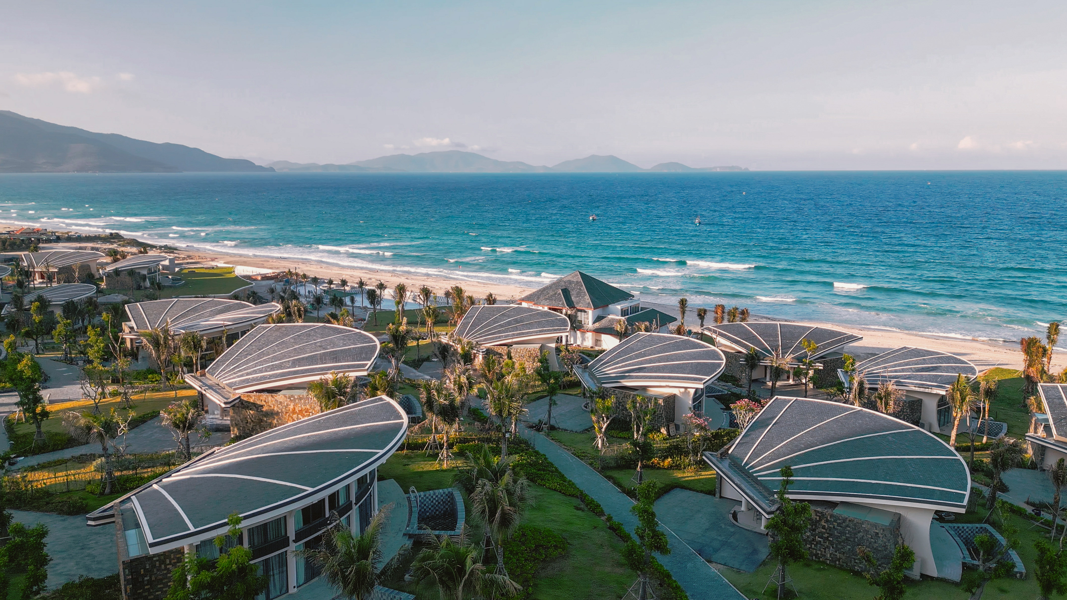 Beachfront resort with unique shell-shaped roofs, turquoise ocean, and mountains.