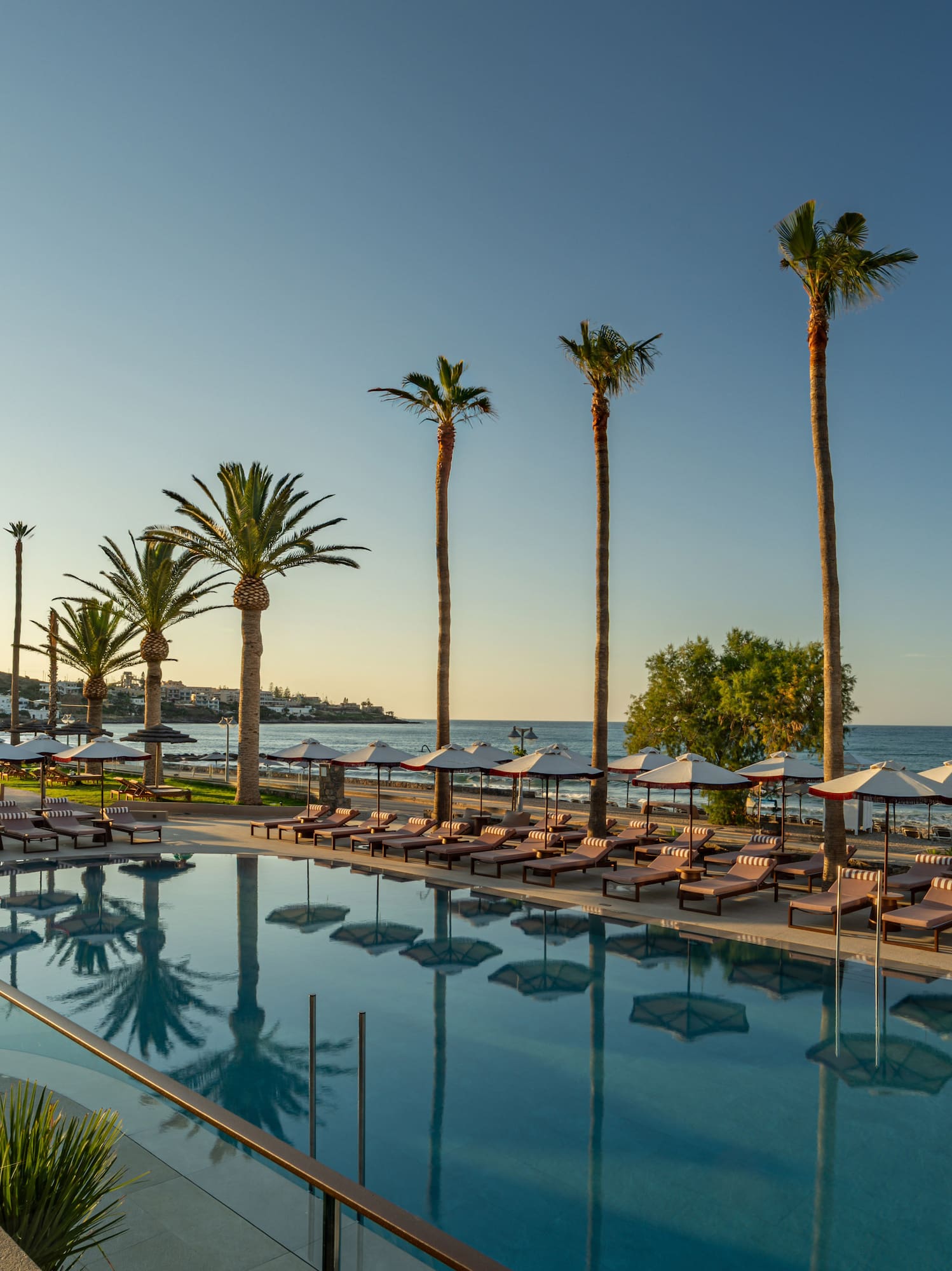 Luxury resort pool with palm trees, sun loungers, beach, and ocean.