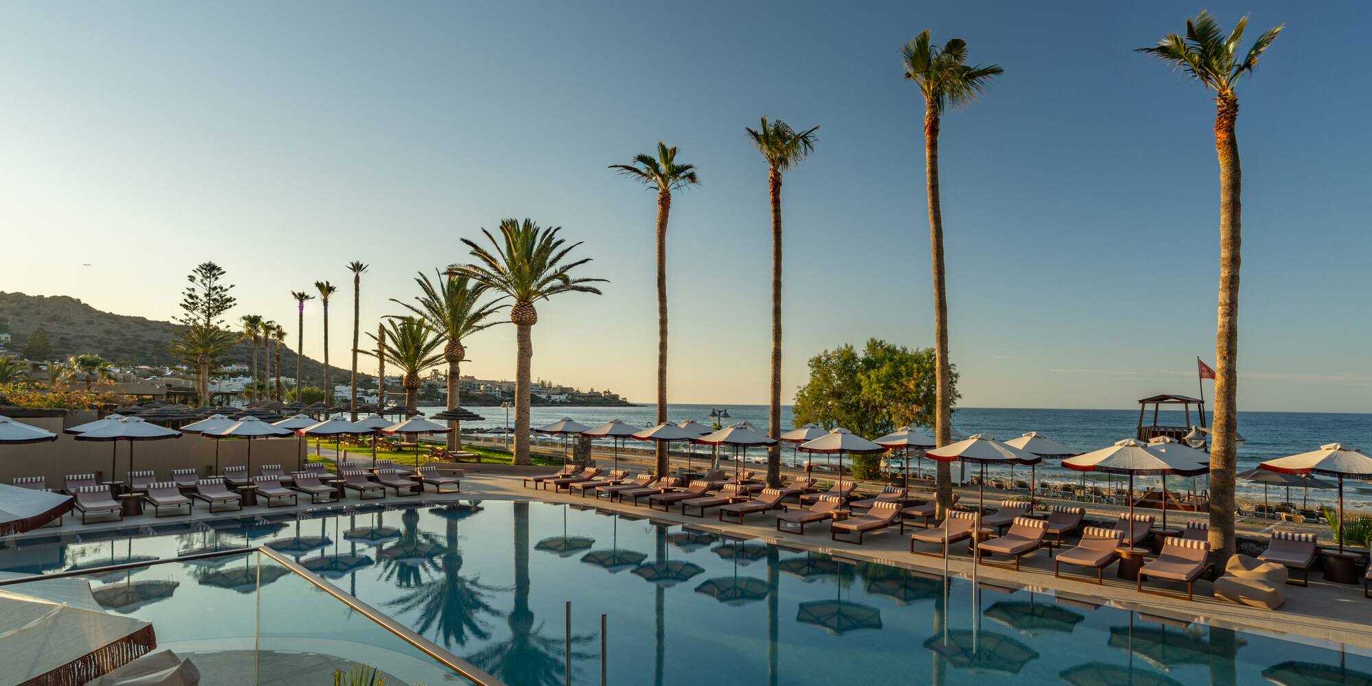 Coastal resort pool with palm trees, sunbeds, beach, and ocean at sunset.