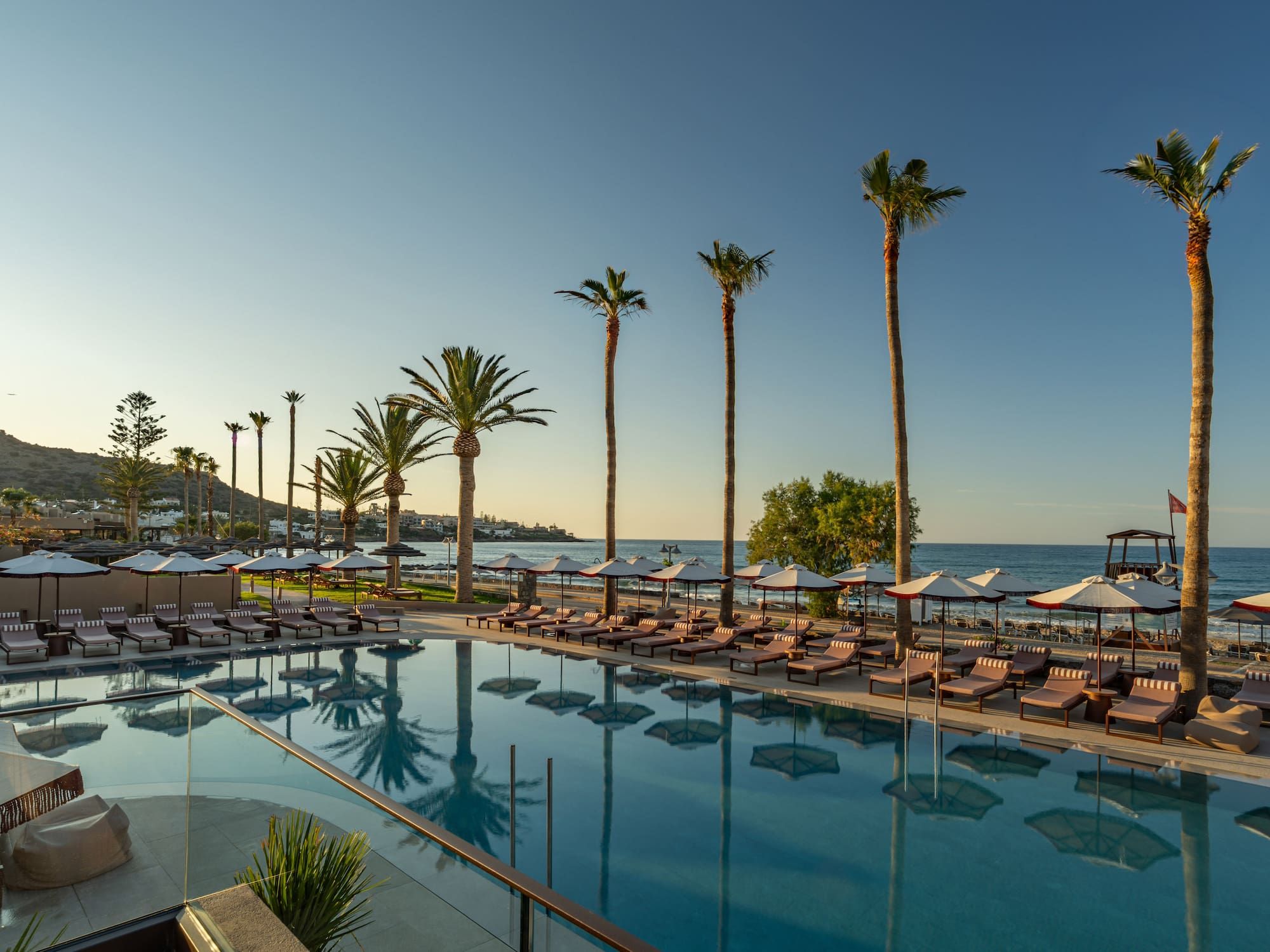 Resort pool with palm trees, sun loungers, and ocean view at sunset.