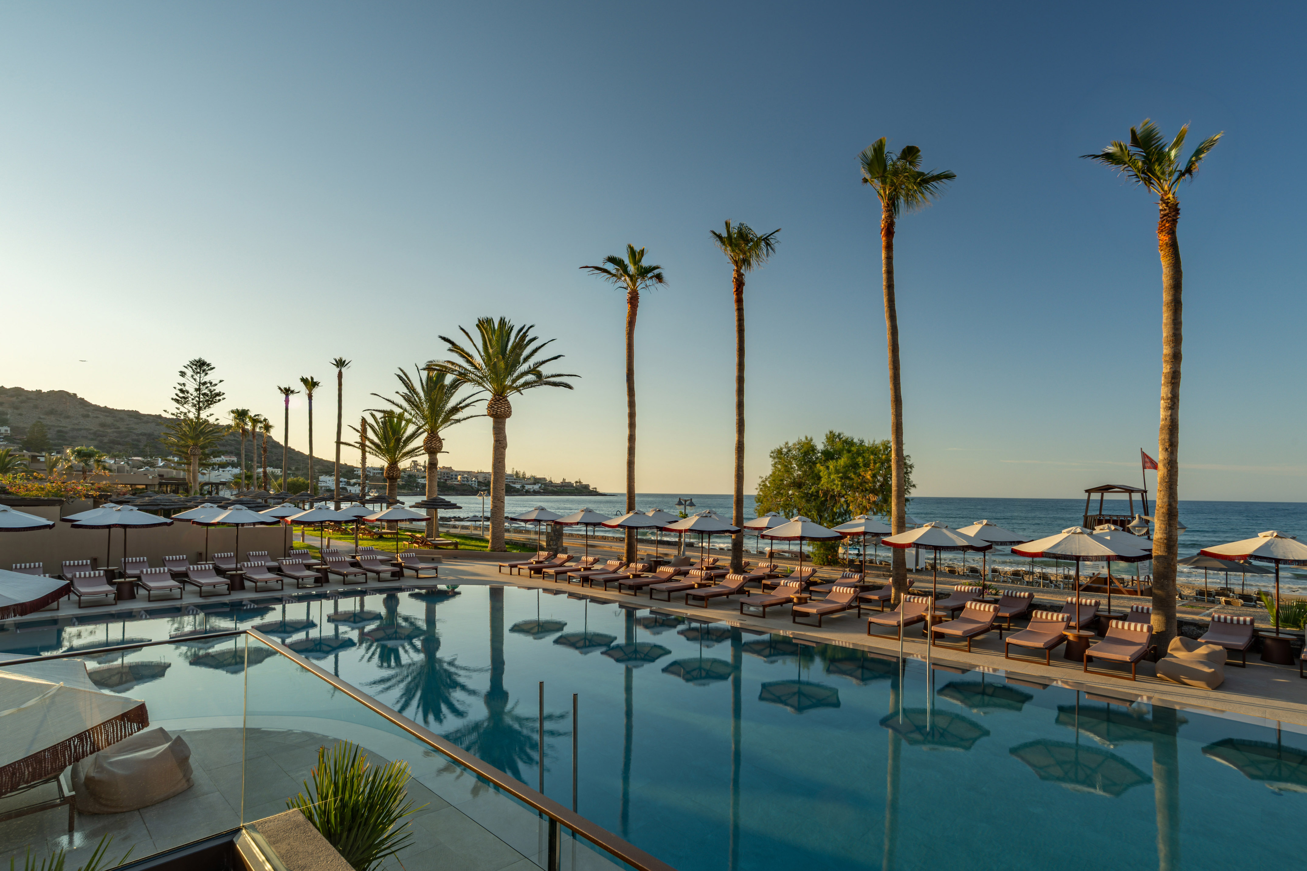 Luxury resort pool with palm trees, sun loungers, beach, and ocean.