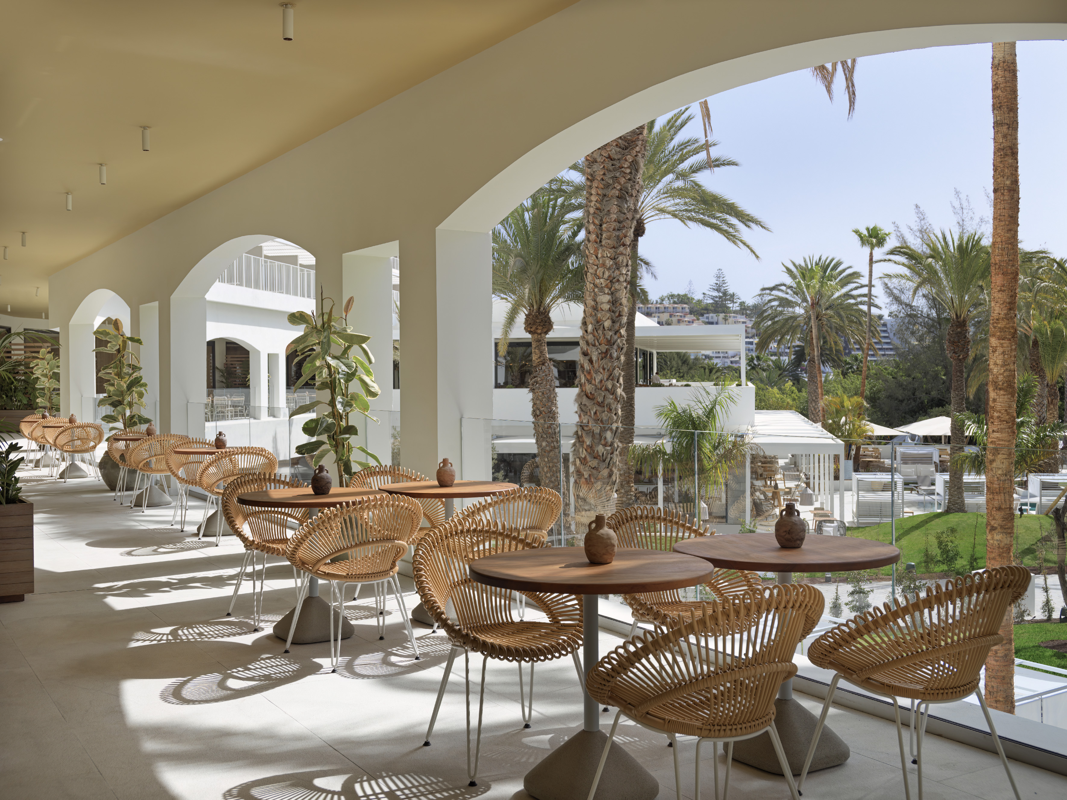 Sunny hotel terrace with wicker dining chairs, round tables, and tropical palm trees.