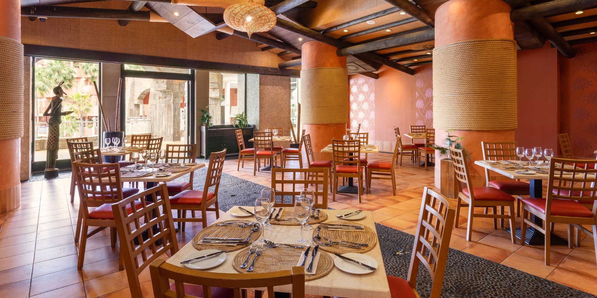 Tropical restaurant dining room with wooden tables, red chairs, rope pillars.