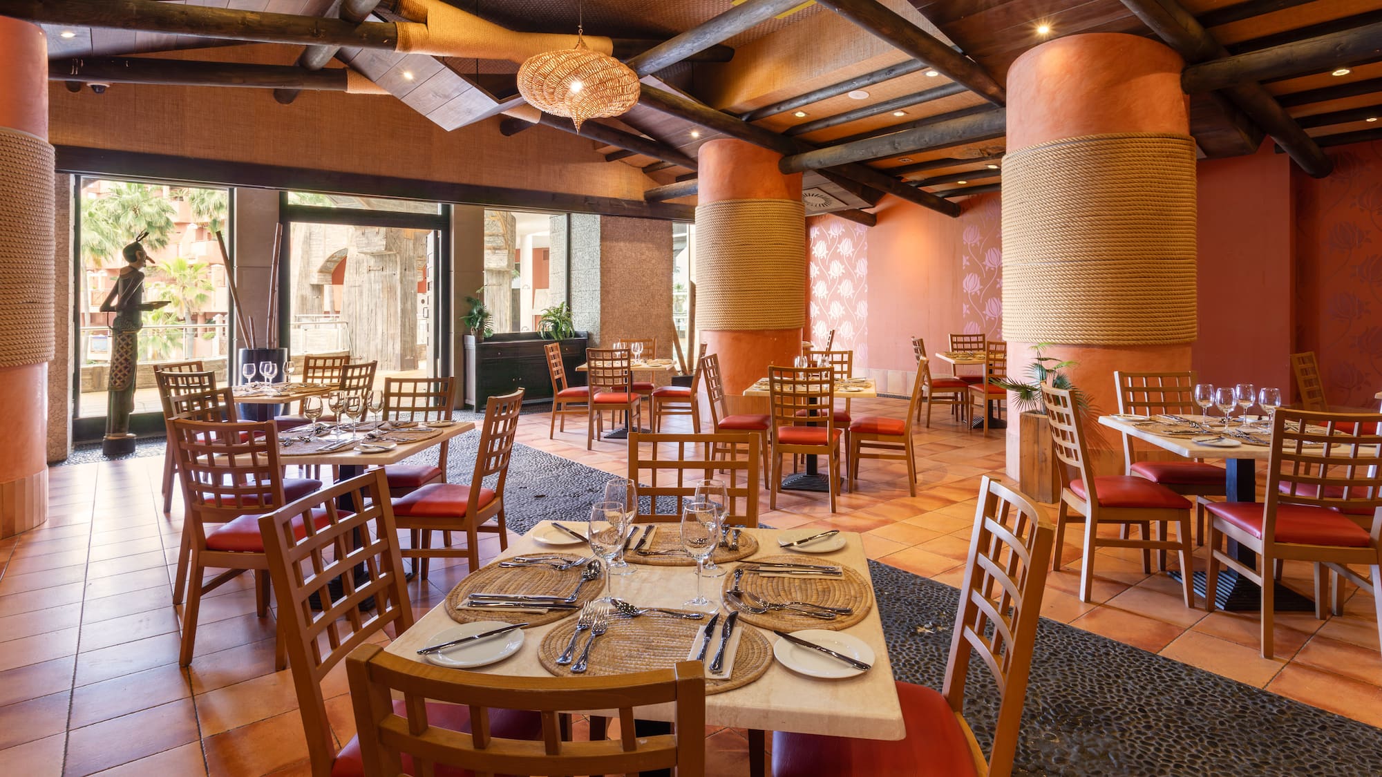 Tropical restaurant dining room with wooden tables, red chairs, rope pillars.