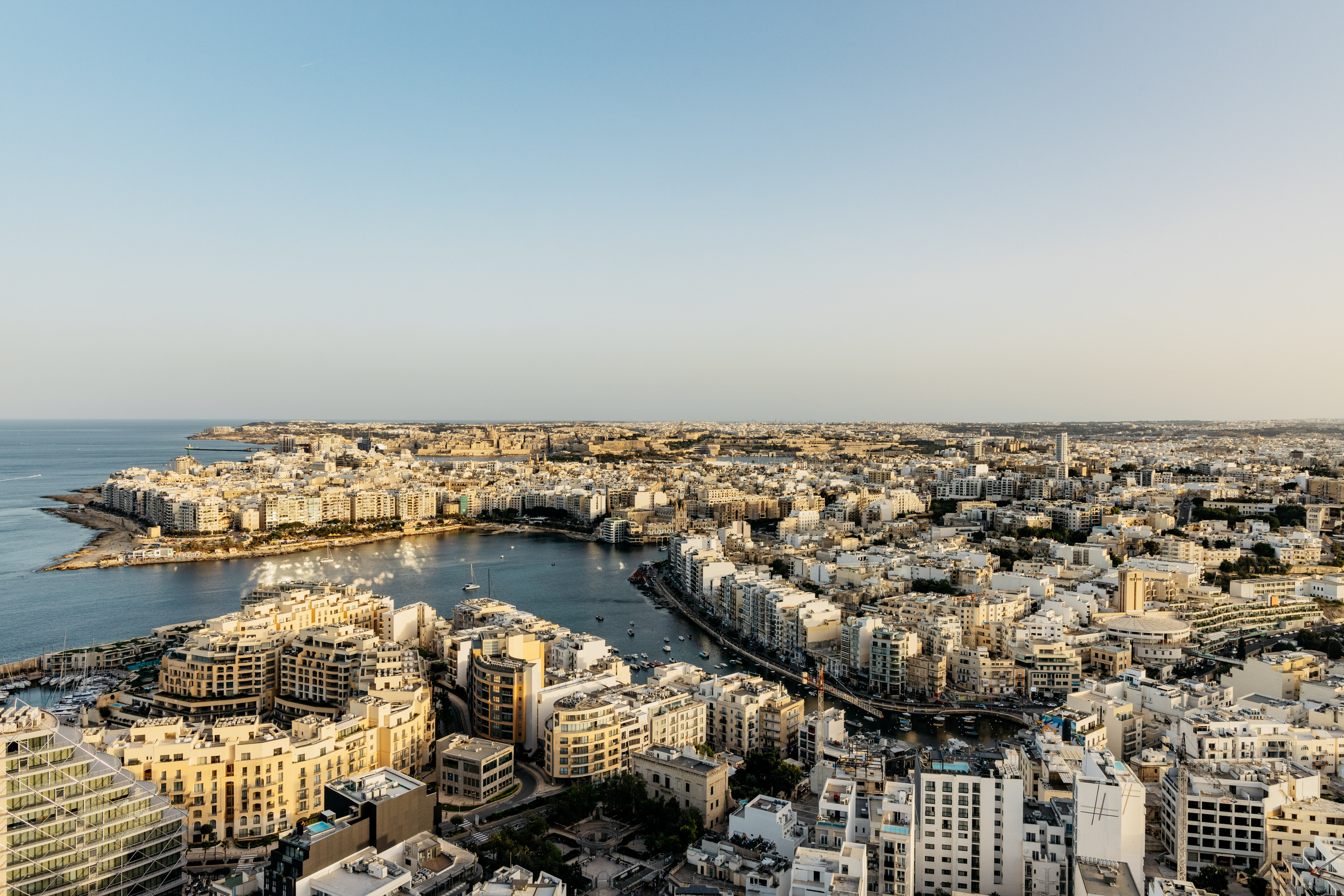Malta coastal city overview: dense buildings line a blue harbor, clear sky.