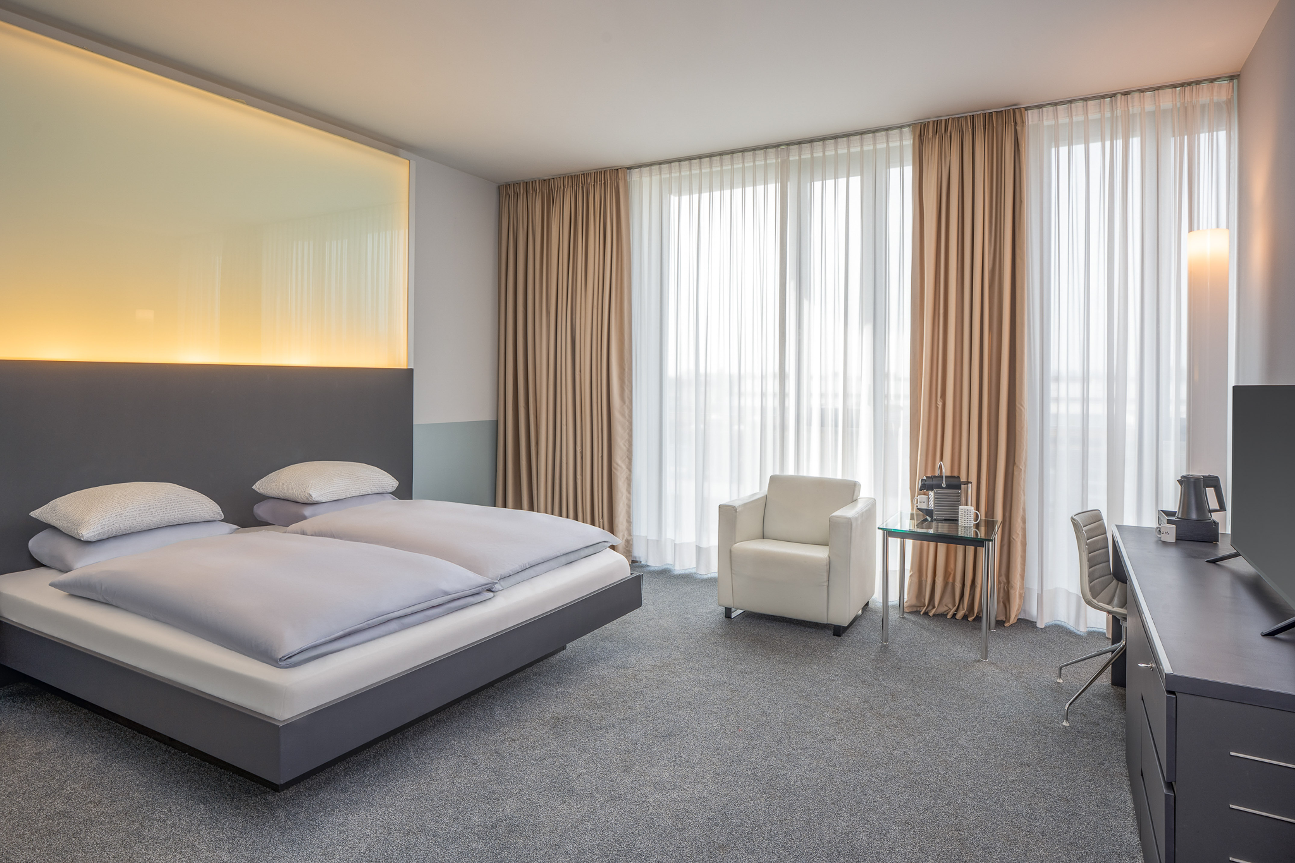 Modern hotel room with grey bed, illuminated headboard, armchair, and window.